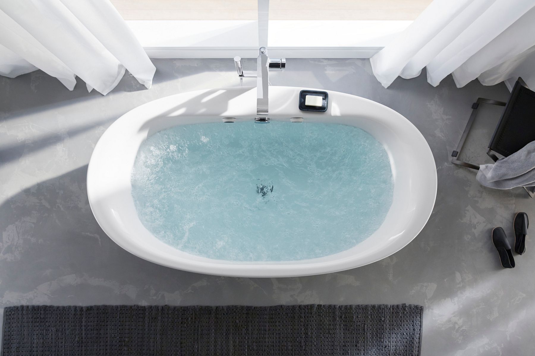 Overhead view of a white freestanding bath filled with water next to a light filled window.