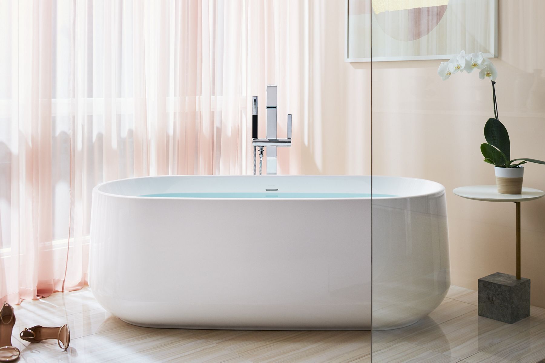 A filled white Ceric bath with a polished chrome bath filler in a room with light colored wood grain flooring and translucent drapes filtering in soft pink light throughout the room. A white orchid sits on a table next to the bath.