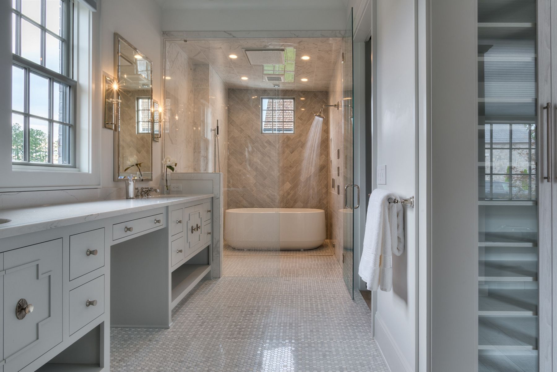 Une salle de bain spacieuse, moderne avec une zone de douche en verre qui comprend un carrelage gris à chevrons et une baignoire ovale autoportante. On aperçoit à l’avant-plan un meuble-lavabo avec un grand miroir, des accessoires élégants et une serviette bien pliée sur un porte-serviette à proximité, créant un cadre serein et luxueux.