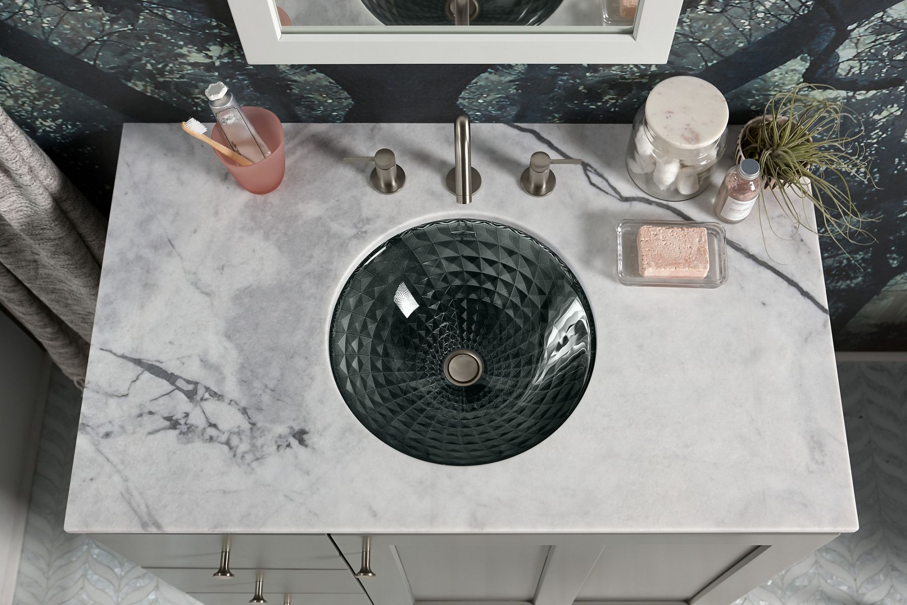 A man uses a cloth and Kohler Faucet Cleaner to clean a mirror in a bathroom with white counters, round bowl sink, and polished chrome single handle faucet.