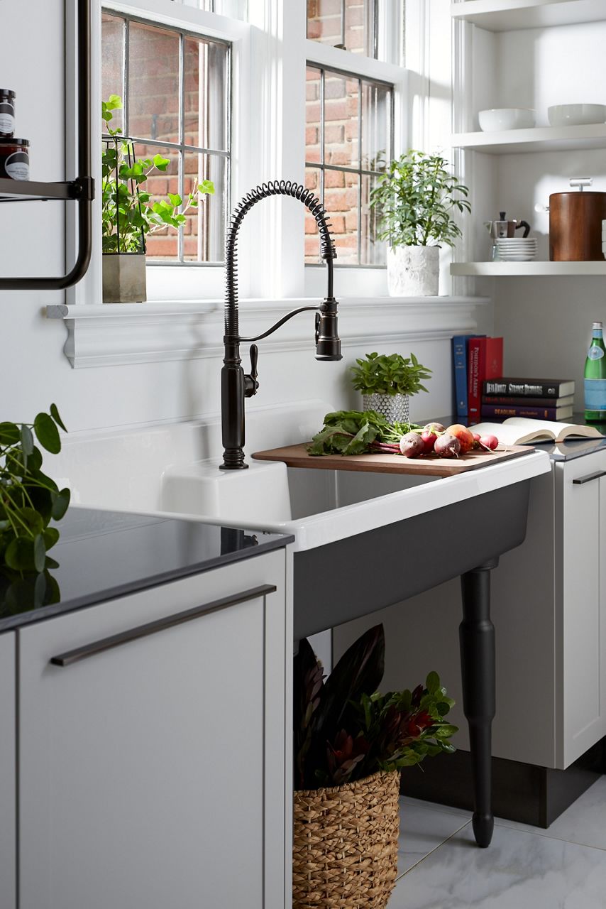 A Tournant semi-professinal faucet in matte black in an enameled cast iron Farmstead sink with legs, in front of a window with plants on the ledge.