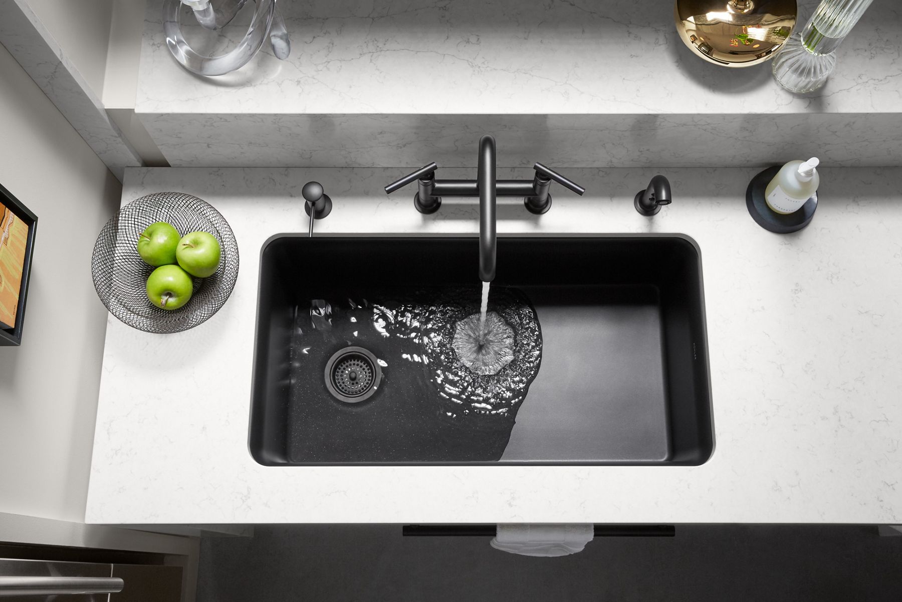 Overhead view of a stone kitchen counter top with a black faucet pouring water into a black sink, with green apples sitting in a glass bowl nearby.