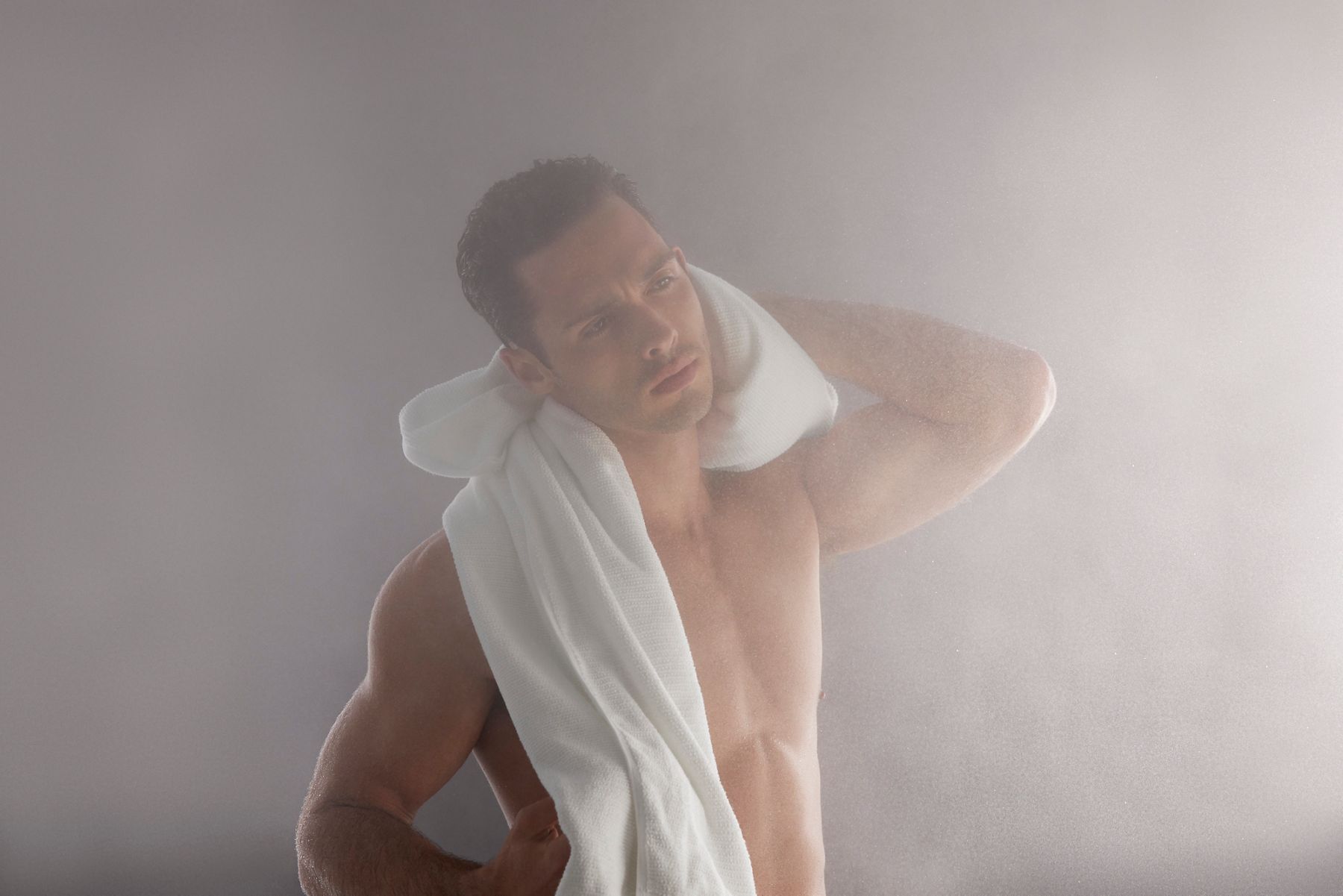 A portrait lifestyle image of a man in a steamy room drying the back of his neck with a white towel that's draped on his shoulder.