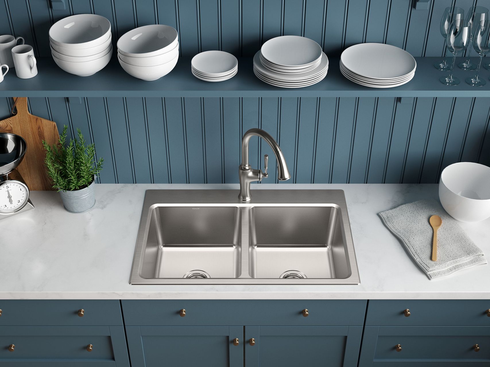 Closeup of stainless steel, double bowl kitchen sink installed in a white countertop with blue bottom cabinets and accent wall with open shelving displaying white bowl and plates.