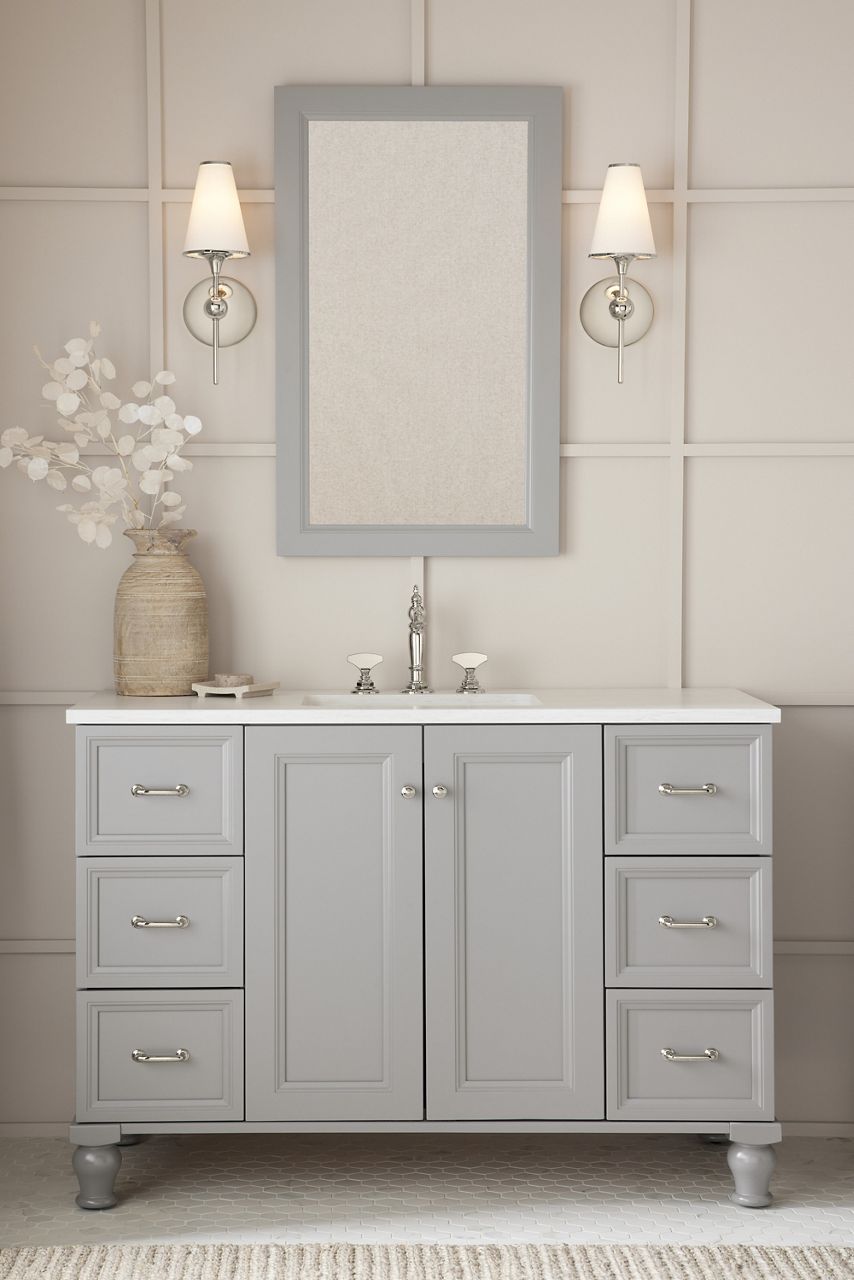 A mohair grey Damask vanity with polished chrome hardware and polished chrome Artifacts faucet, along with a mirror and two sconce lights in a bathrooom with off-white walls and raised square framing.