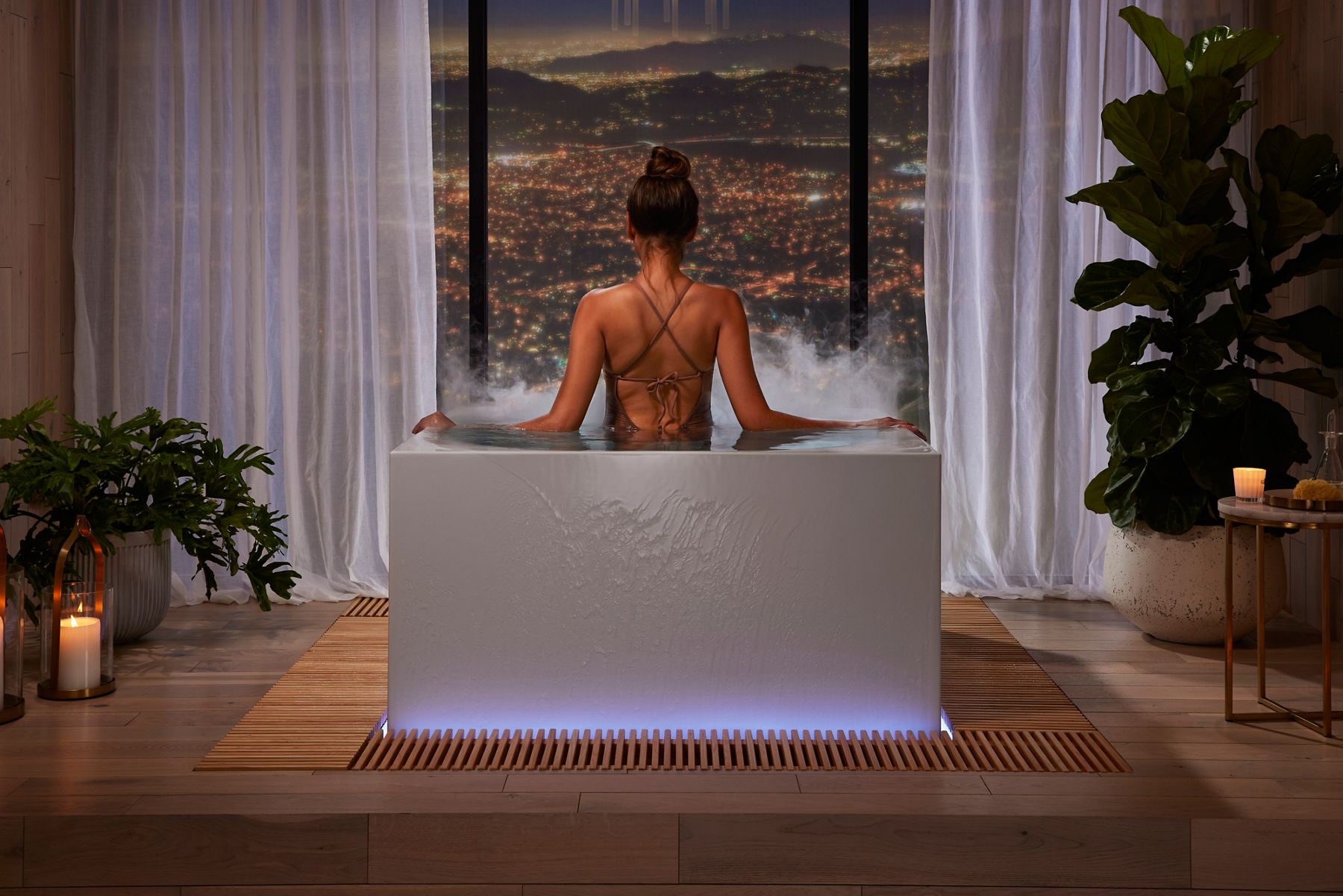 Woman sitting in a bathtub facing large windows with a scenic city view, surrounded by candles and plants.