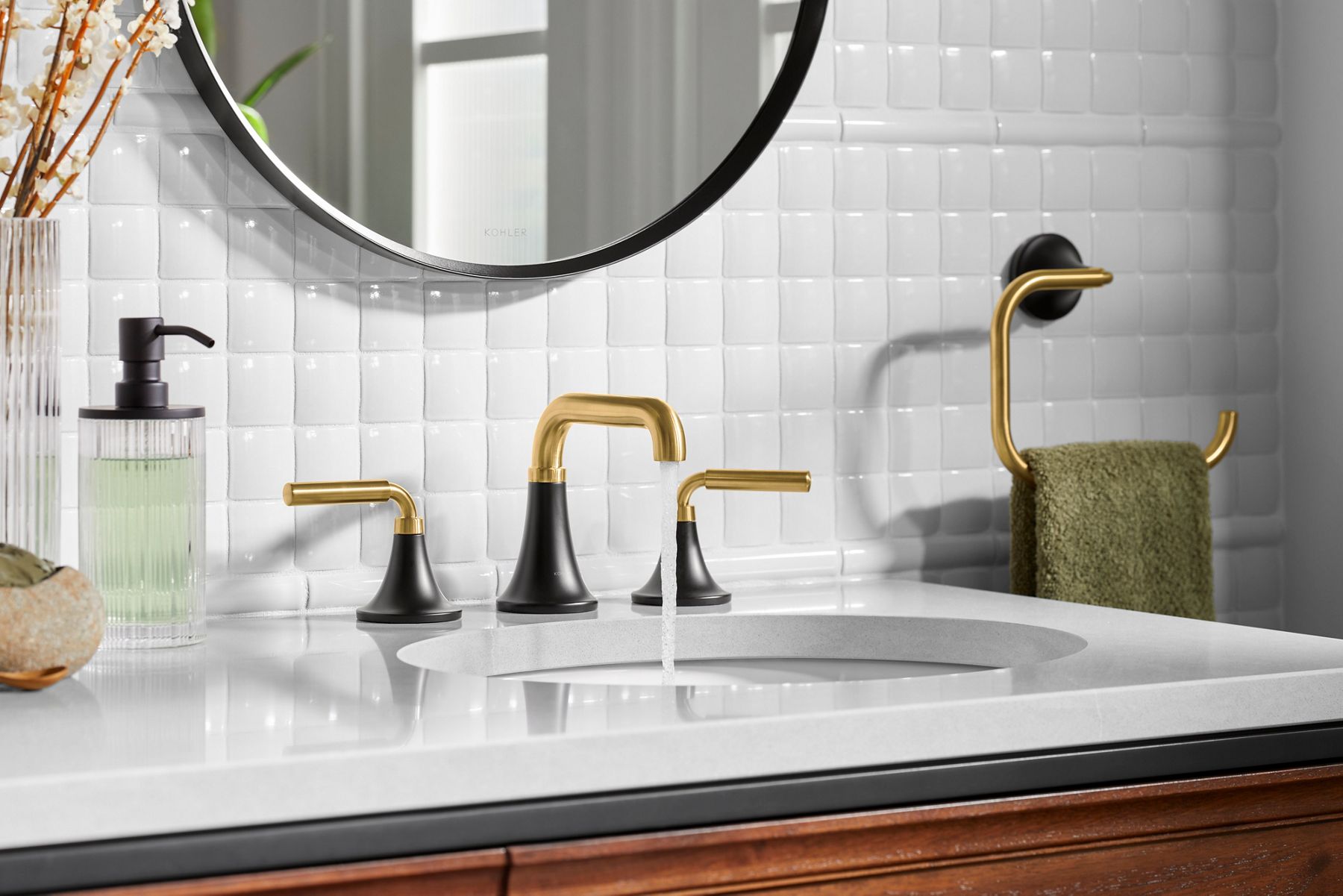 A two-tone matte black and brushed brass widespread faucet with a tapered base on a thie counter top and glossy white tile backdrop.