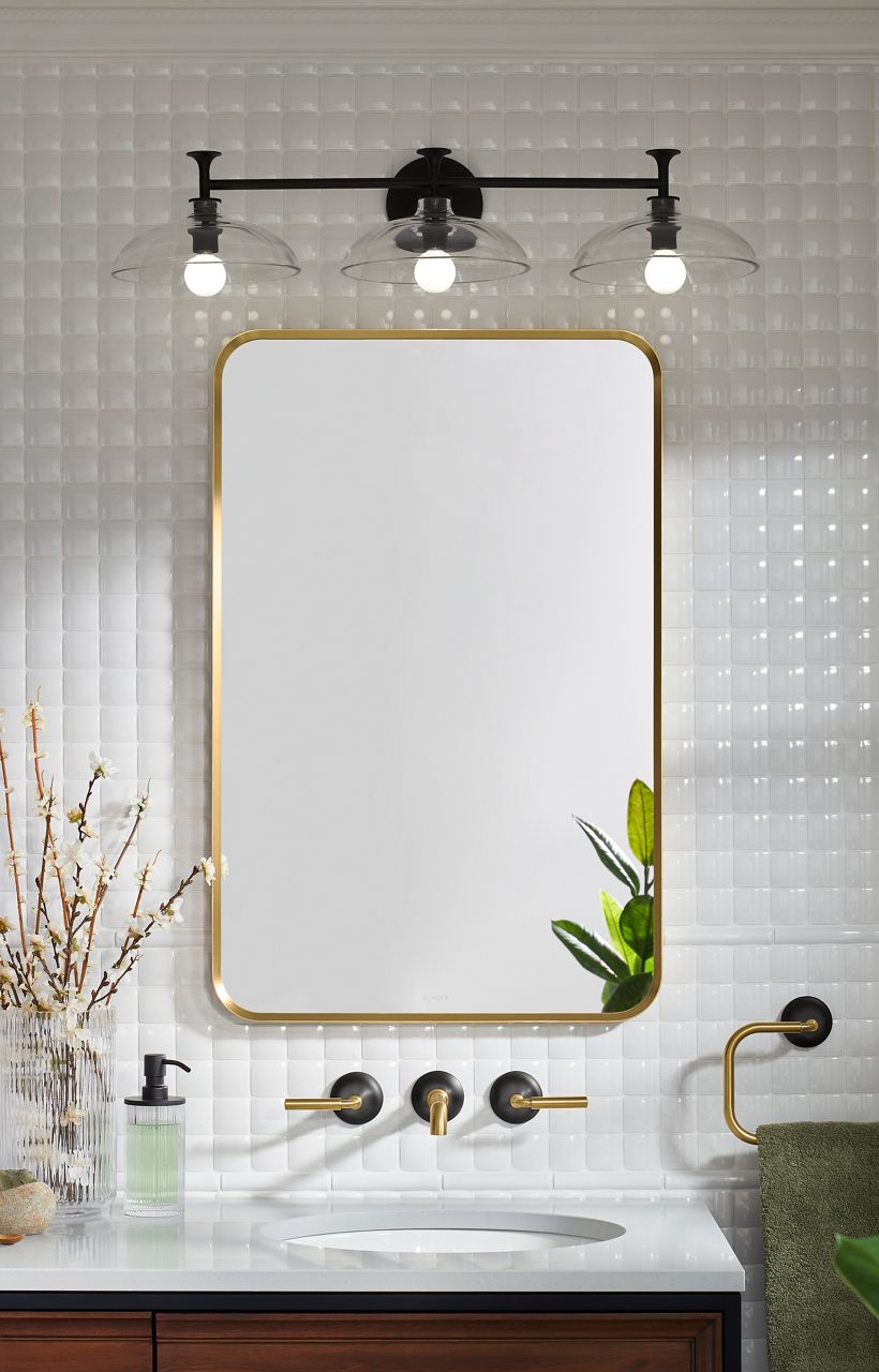 A brass-framed rectangular mirror on a glossy white tile wall with a three light sconce above it.