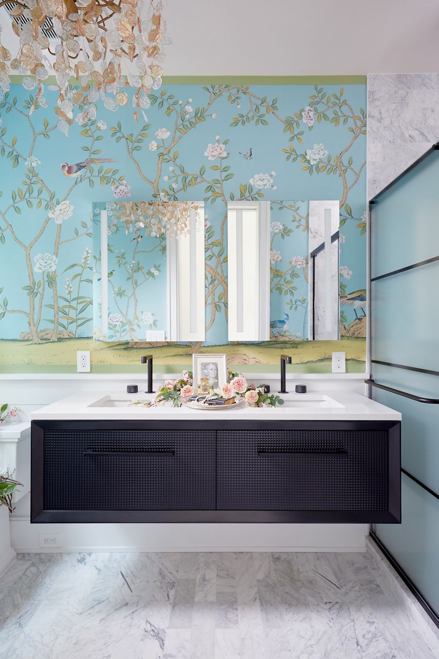 A bathroom styling space with a black wall-mount double vanity with white counter top and undermount sinks, black faucets, rectangular frameless mirrors and wallpaper of light blue with a nature scene print of trees and flowers.