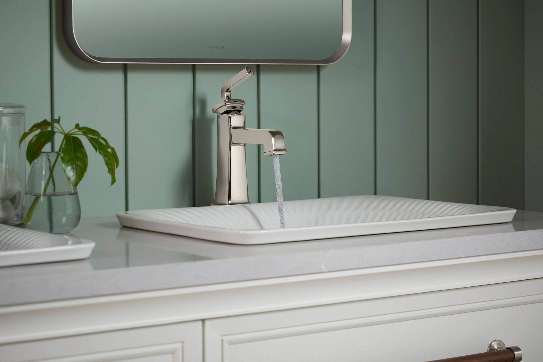 A square faucet pouring water into a scalloped white sink in a white vanity.