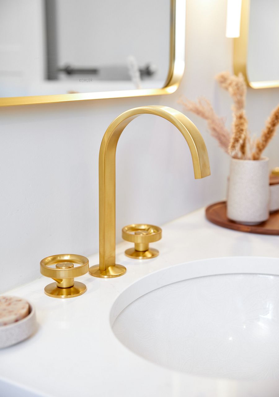 A sleek contemporary bathroom faucet in brushed moderne brass on a white counter, above a white sink, with a mirror framed in brushed moderne brass above it.