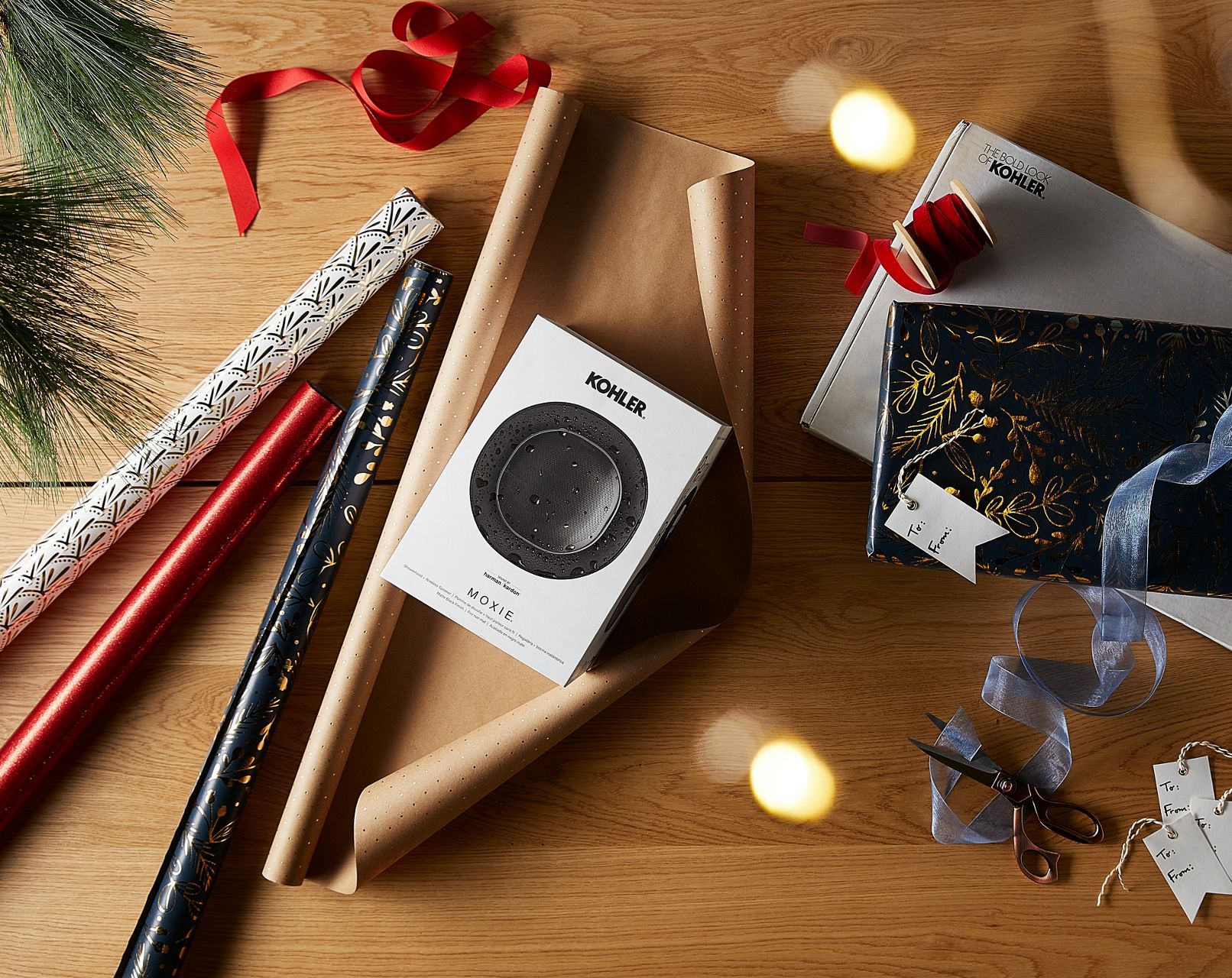 Looking down at a table of wrapping paper rolls and ribbon, there is a KOHLER Moxie showerhead package that is about to be wrapped for a gift.