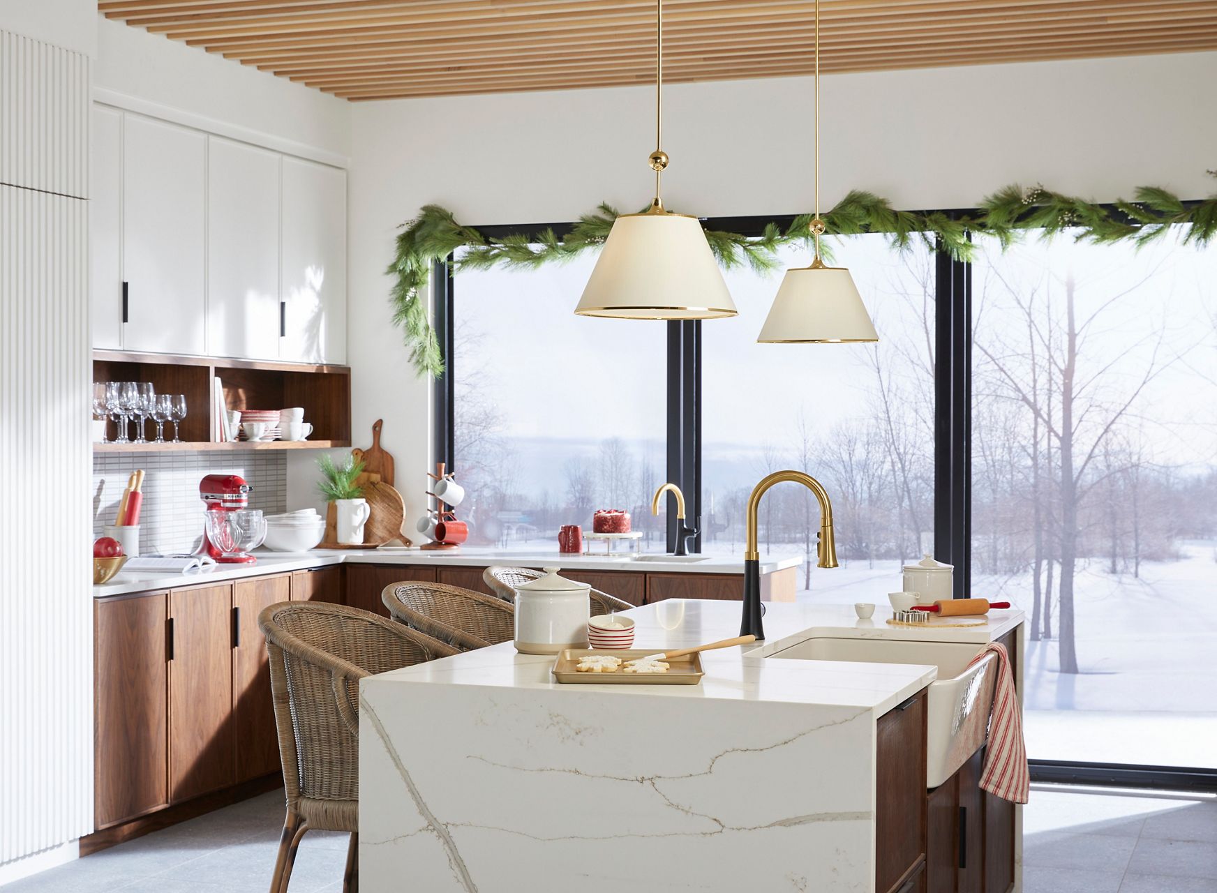 Plan large d’une cuisine décorée de verdure festive et de décorations de vacances d’hiver. Il y a des biscuits sur le grand comptoir de l’îlot, où un évier est centré et un grand robinet deux tons noir mat et laiton est monté. Un évier de bar plus petit et un robinet de bar deux tons plus petit se trouvent à l’arrière-plan, près d’un mur extérieur de fenêtres.
