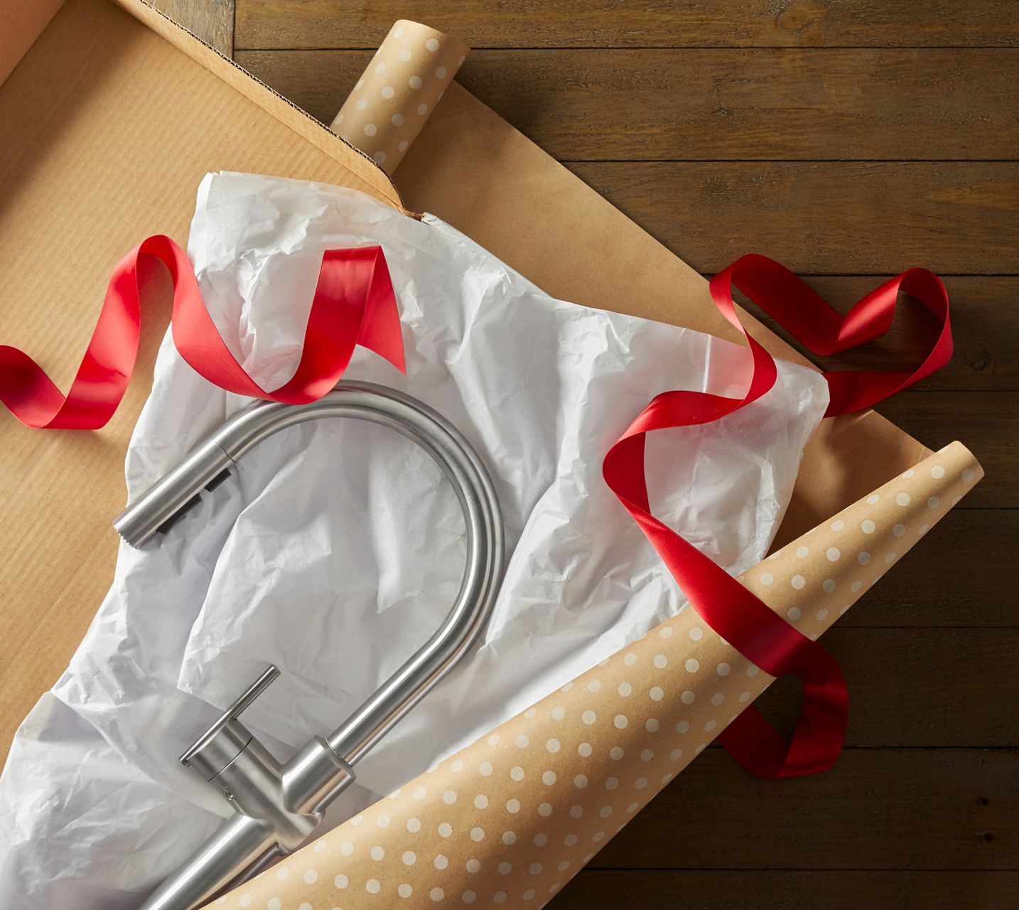 A polished chrome kitchen faucet is placed on a sheet of tissue paper, laid over a roll of holiday gift wrap and ribbons, about to be wrapped as a present.