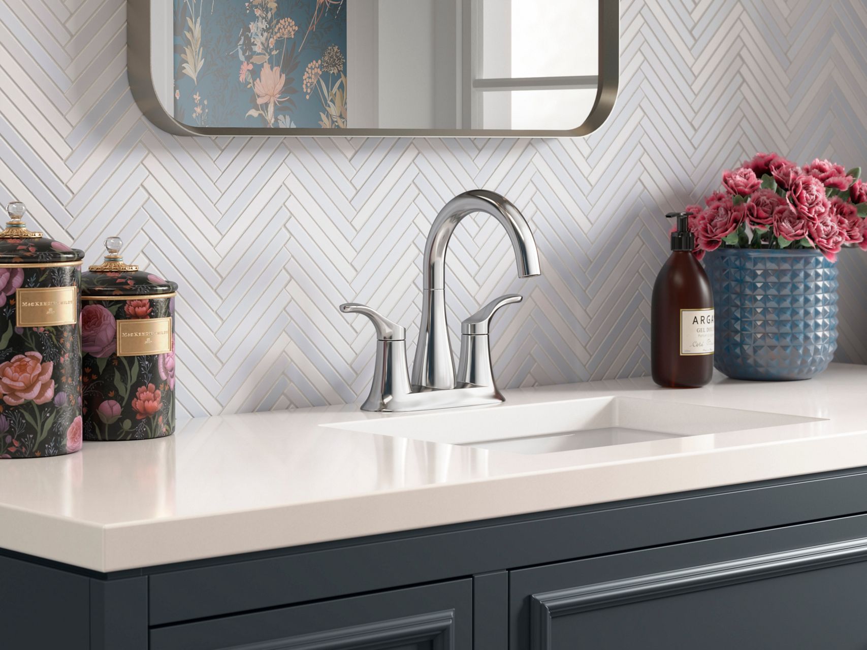 A chrome centerset faucet with soft, flowing lines on an off-white counter top with a wall of thin light gray tile in a herringbone pattern.
