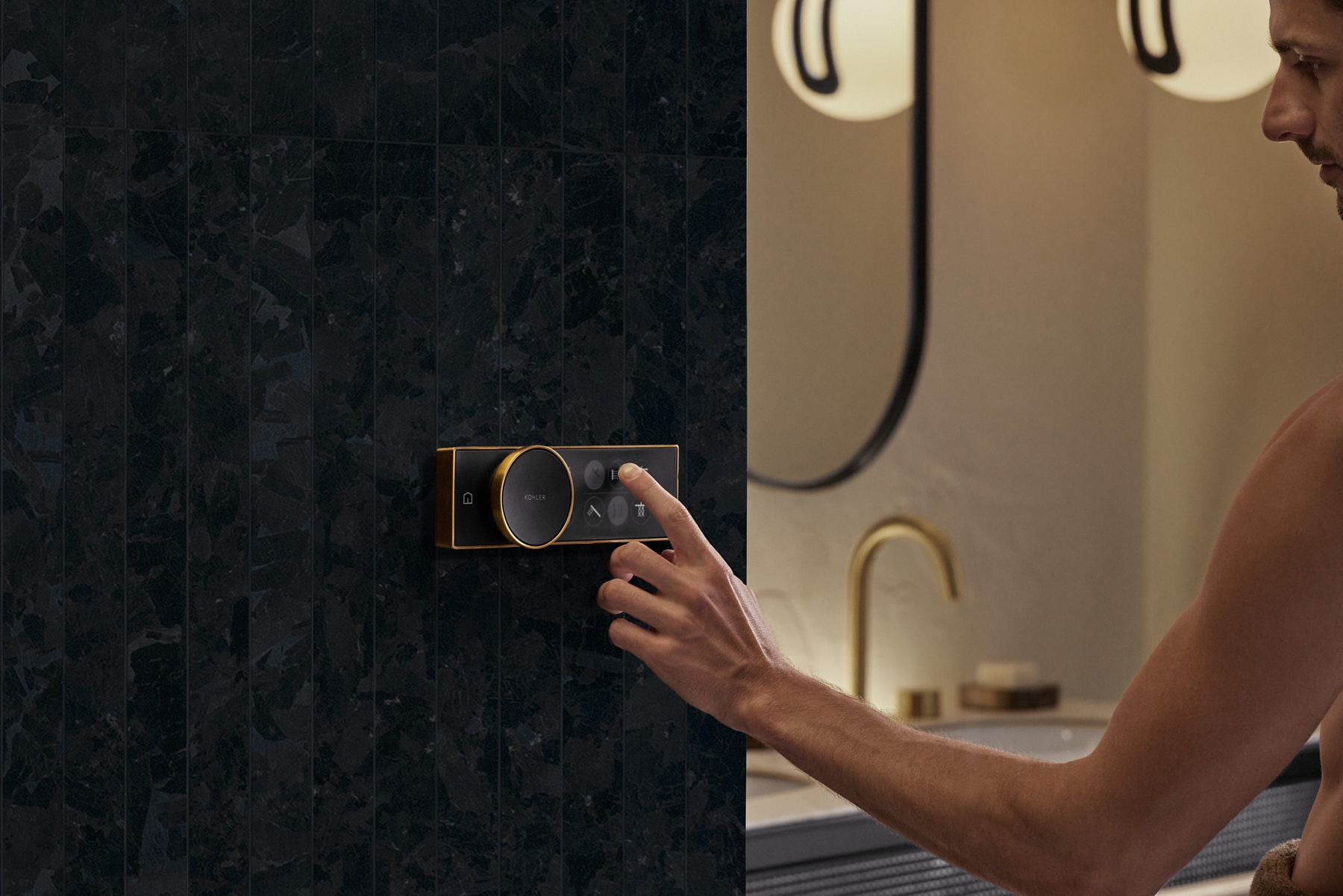 Une main tend la main pour toucher un module de commande de douche Anthem noir avec une robinetterie dorée contre un mur en carreaux de pierre gris foncé.