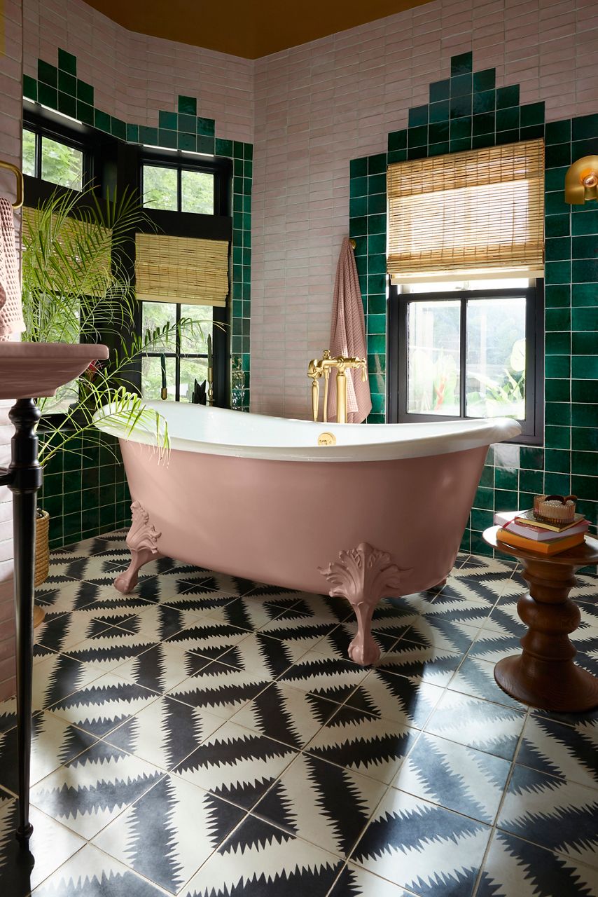 Freestanding clawfoot bathtub in a colorful bathroom featuring green tile walls, patterned black-and-white flooring, and large windows with natural light.