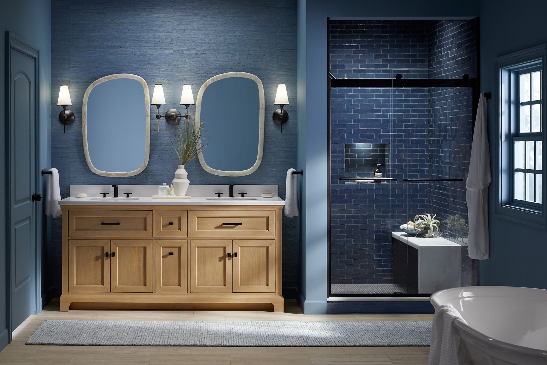 Modern double-sink wood vanity with black hardware, white countertop, and two oval mirrors, set against a navy blue textured wall with wall scones and a walk-in shower on the right.