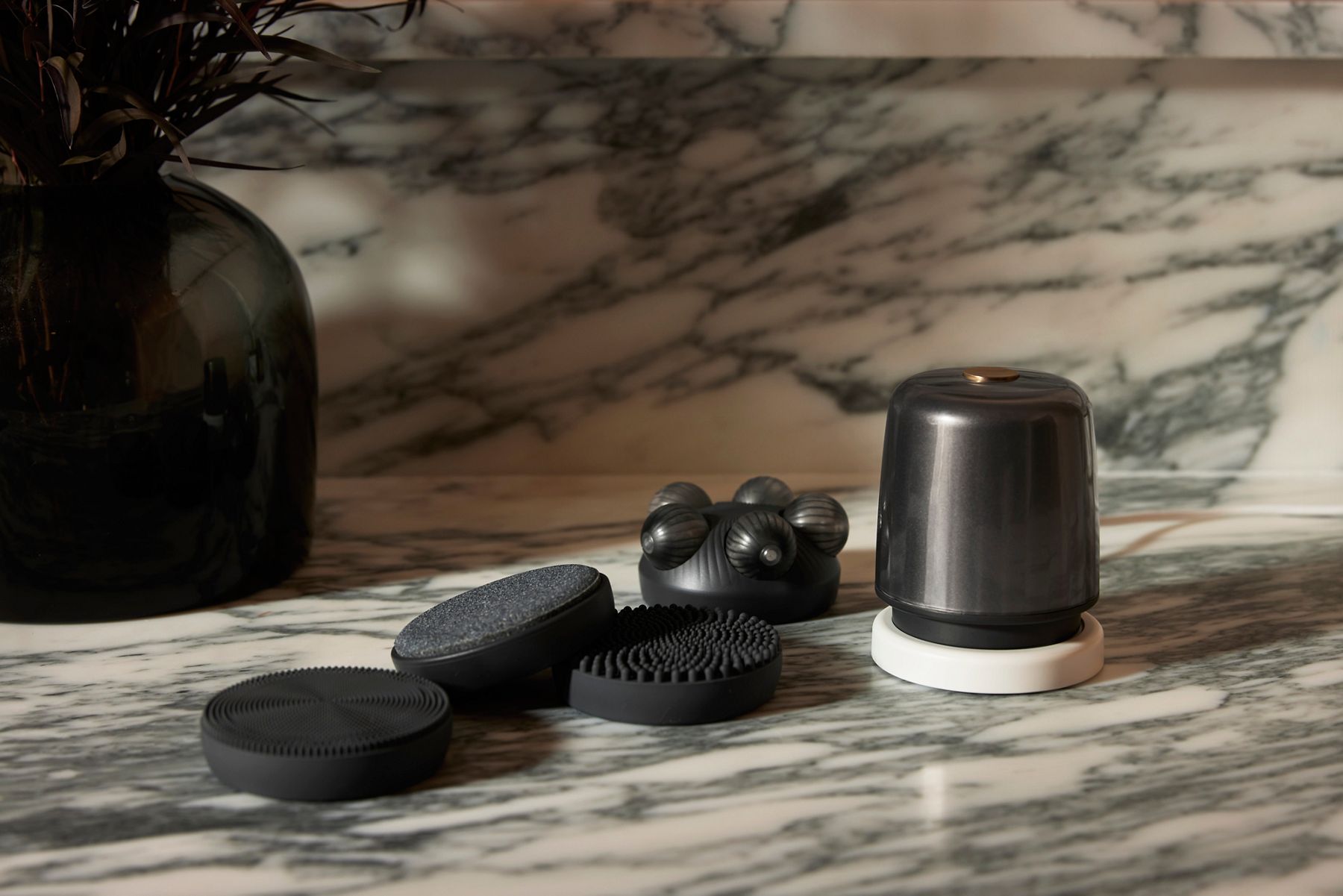 A portrait lifestyle image of bath products placed on a marble counter. The products include a black body massager and interchangeable attachments.