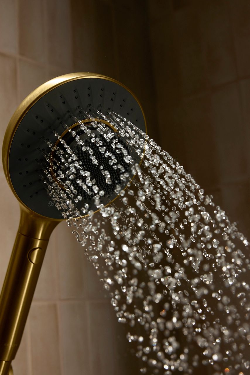 A close-up portrait product image of a gold and black running shower head attached to a beige tiled wall.