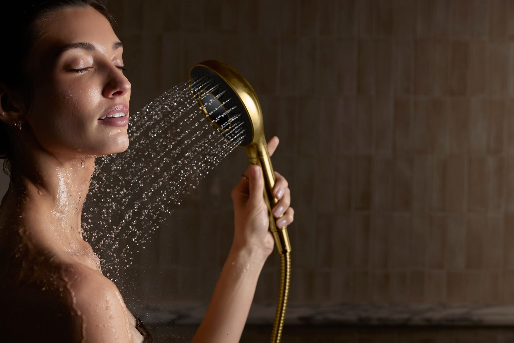 A close-up portrait shot of a woman using a gold-toned shower head. The shower head is placed on her neck with water running.