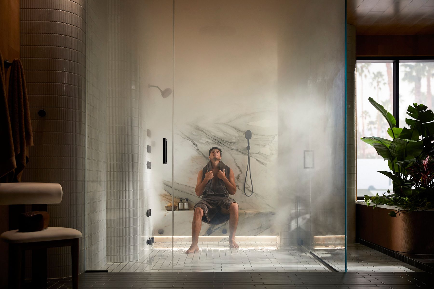 A man sitting in a glass-walled steam shower, wearing swim trunks and holding a towel.