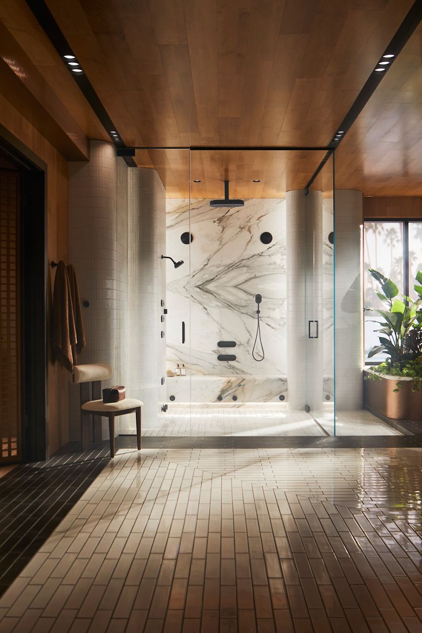 A spacious, luxury shower with a glass enclosure, featuring marble walls, black shower fixtures, a built-in bench, and an overhead rain showerhead. The shower area is complemented by a wooden ceiling, light-colored tiled floor, and a small table with a plant visible outside.