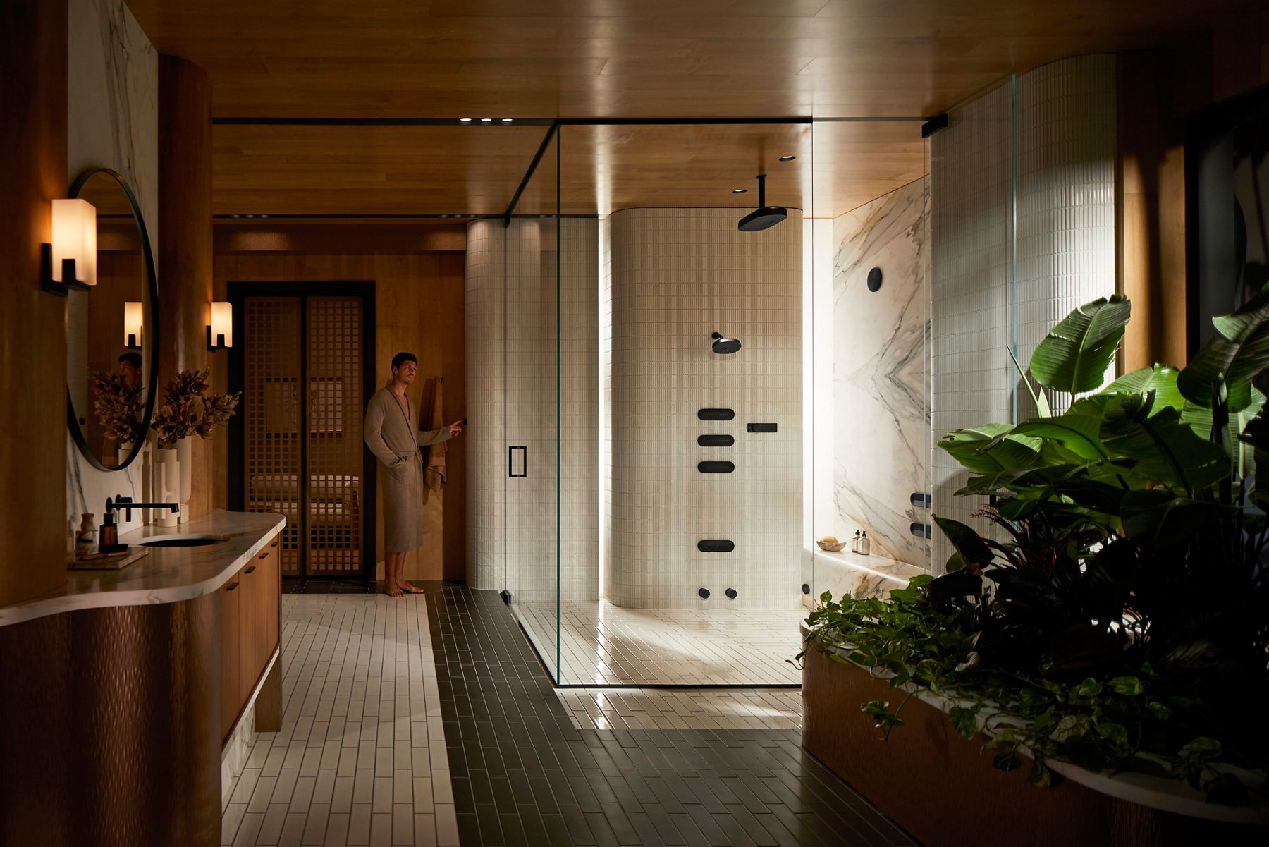 A modern, glass-enclosed shower with sleek black fixtures, including an overhead rain showerhead and multiple wall-mounted controls. The space features marble accents, a built-in bench, and warm lighting, with a green plant visible in the foreground.