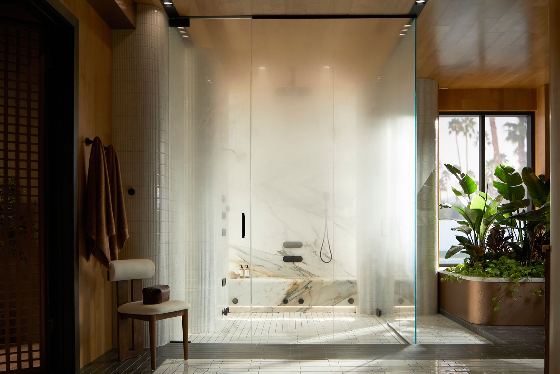 A modern glass-enclosed shower with a sleek and minimalistic design, featuring a built-in bench, wall-mounted fixtures, and soft lighting, surrounded by light-colored tiles and a nearby plant outside the shower area.