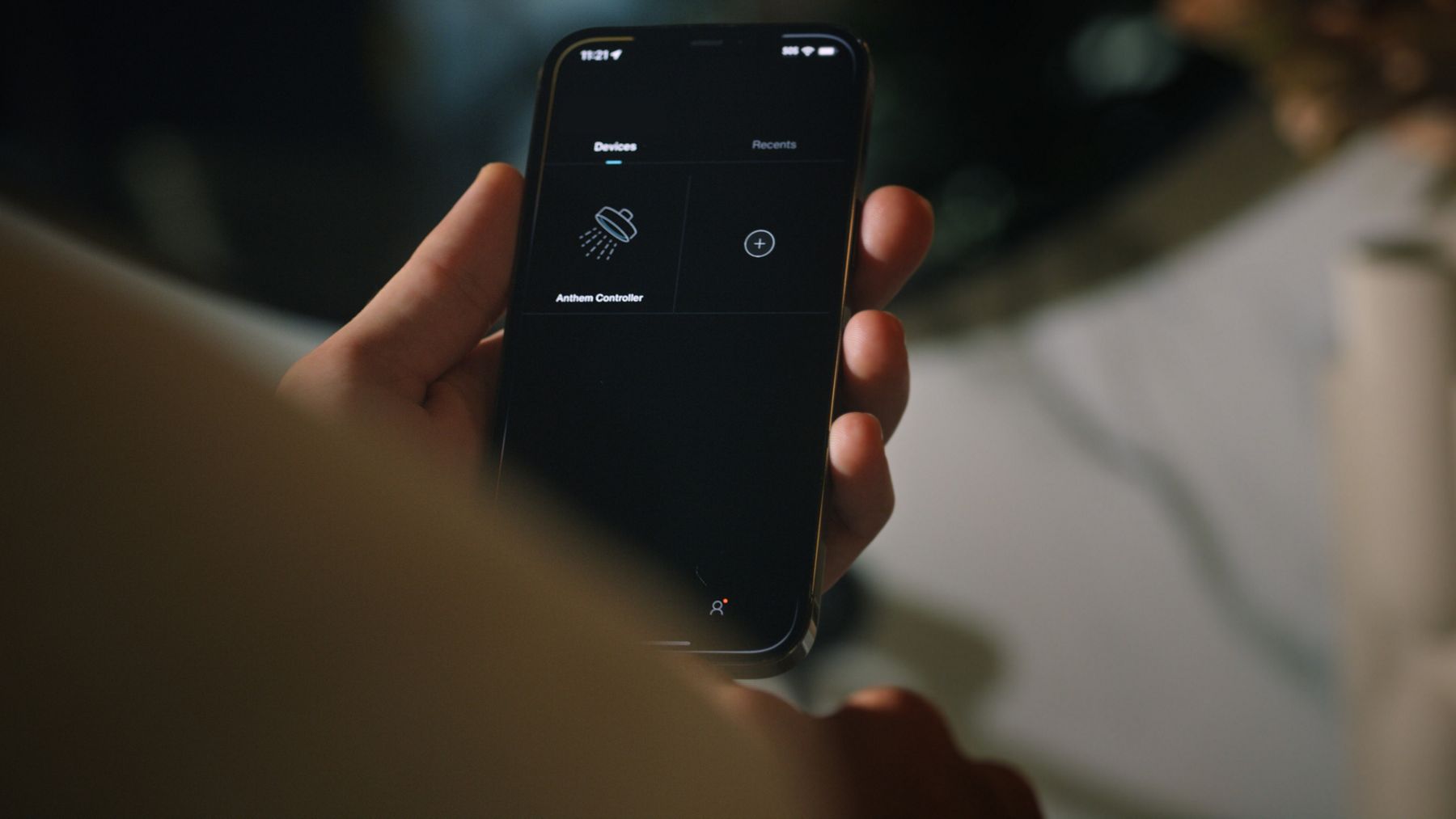 A person holding a smartphone displaying a dark-themed app interface with icons for shower settings and lighting control. The screen shows options to customize the shower experience, with minimalistic white icons on a black background.