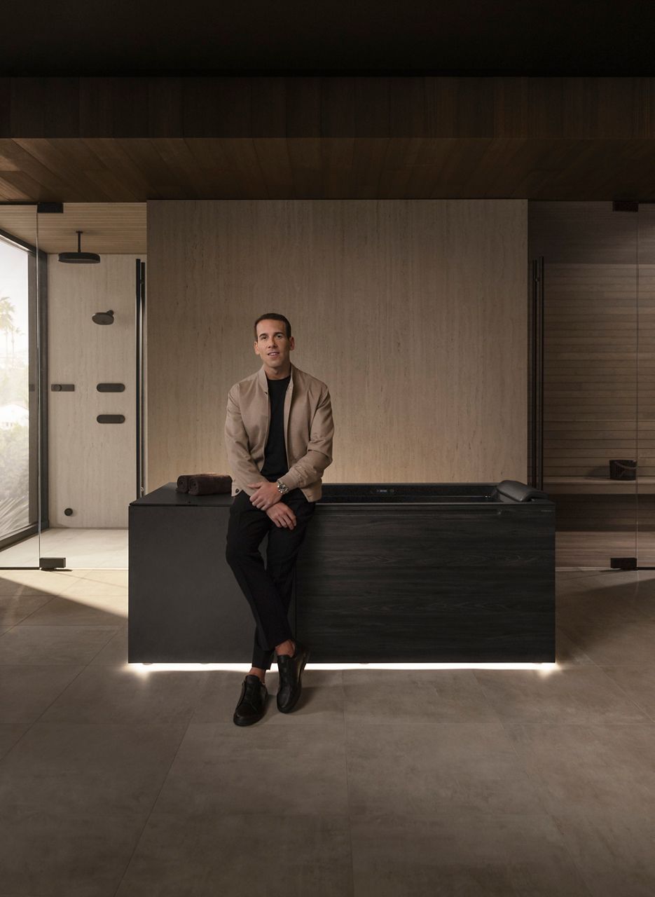 Dr. Jonathan Leary, founder of Remedy Place, sitting on a modern dark wooden ice bath in a luxurious, minimalist bathroom with soft lighting.