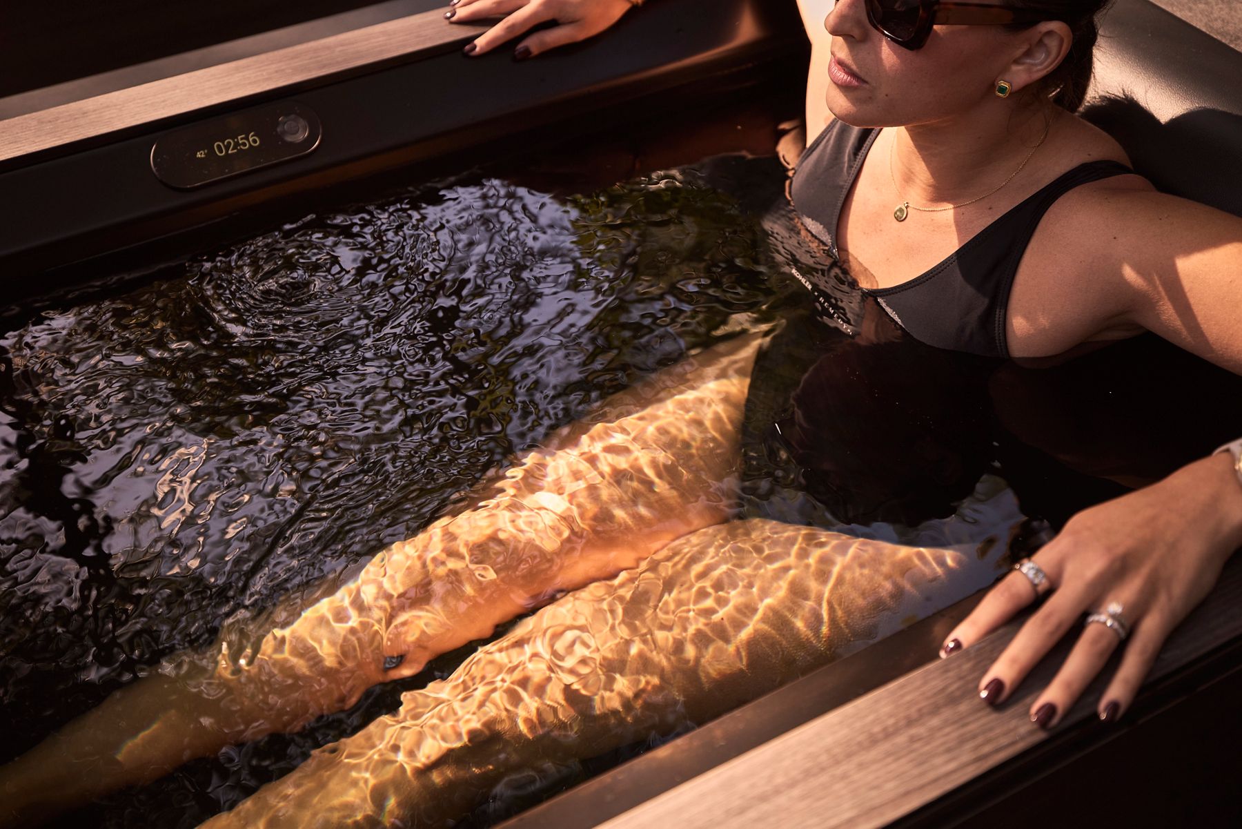 A woman wearing dark sunglasses and a black swimsuit relaxing in a ice bath.