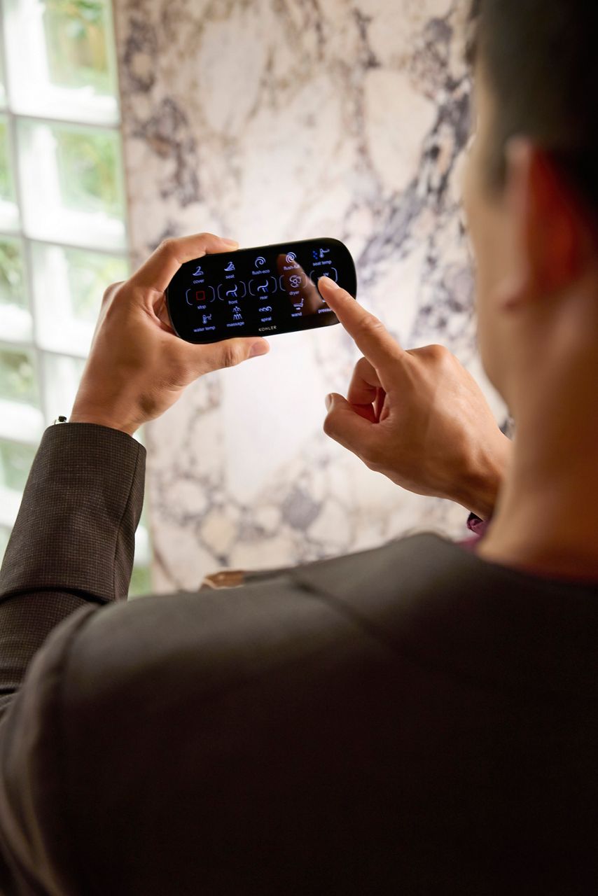 A person uses a touch-screen remote control in a bright, gray bathroom.