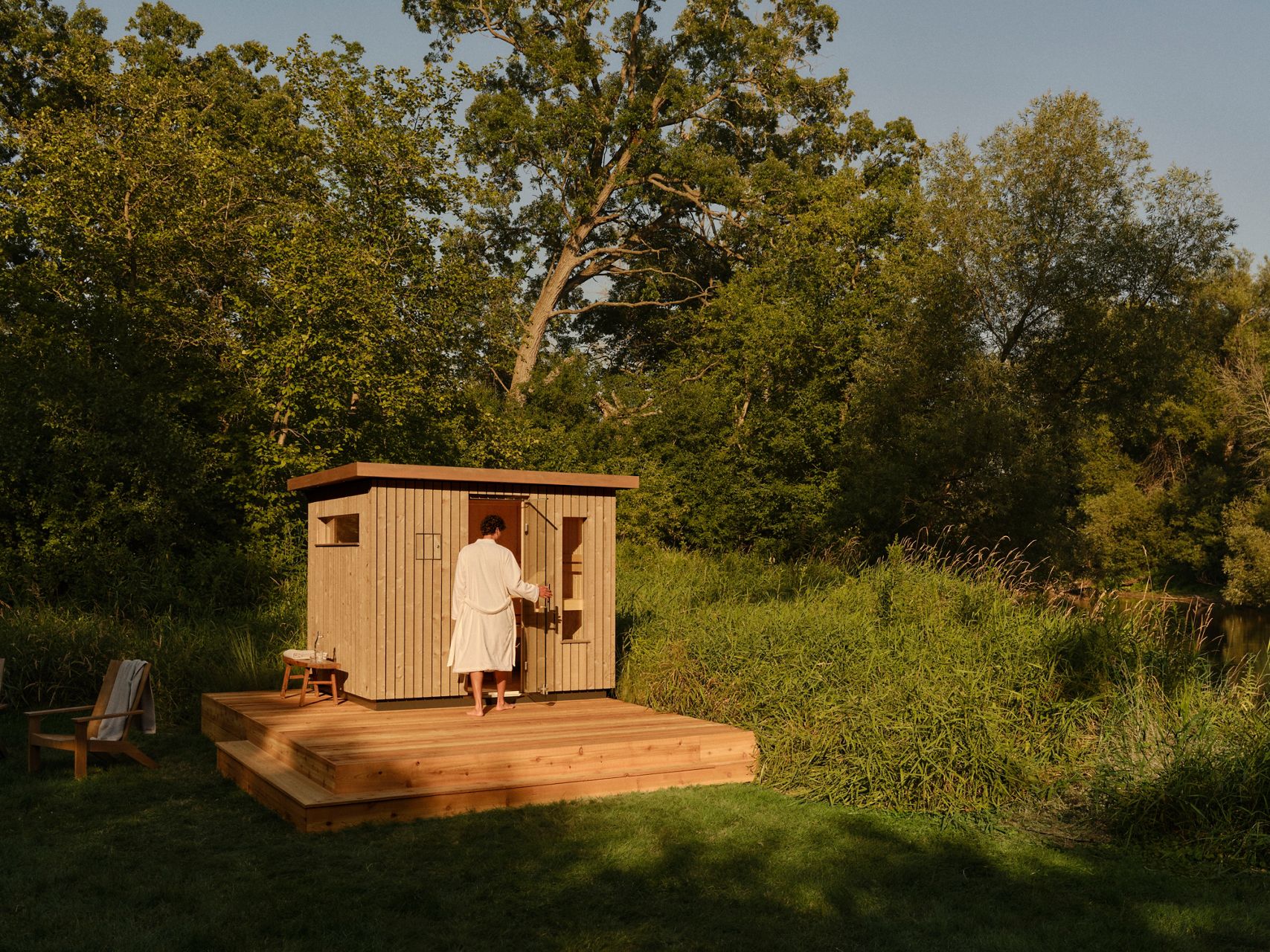 Person walking toward a small outdoor wooden sauna surrounded by greenery.