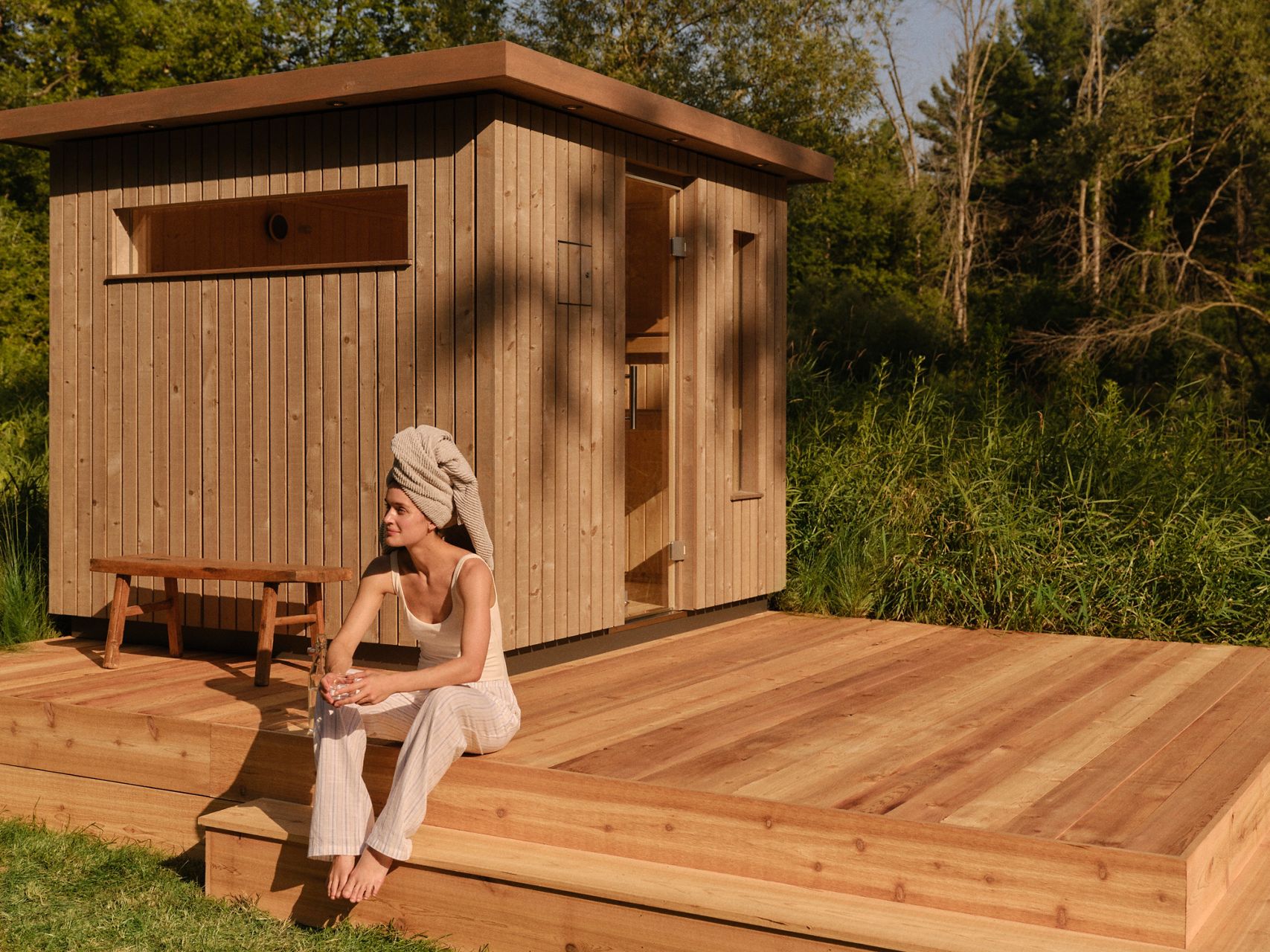 A person wearing light-colored clothing sits on a wooden deck beside a modern wooden outdoor sauna, surrounded by greenery and trees on a sunny day.