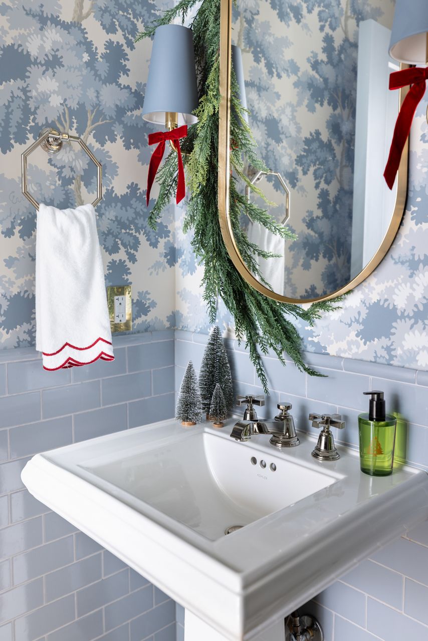 Jenn Lake’s bathroom with floral wallpaper, pedestal sink, and gold-framed mirror decorated with Christmas garland.