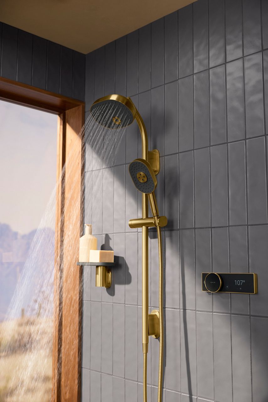 KOHLER Anthem EvoCycle shower system in a gold finish, featuring an overhead showerhead and hand shower mounted on a dark gray tiled wall beside a window.