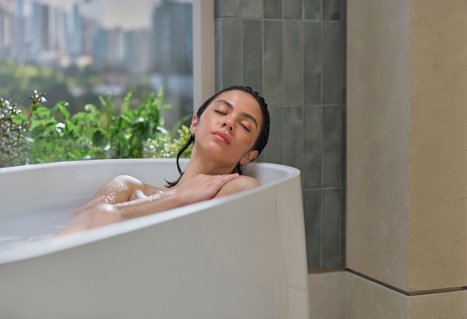 Person relaxing in a white freestanding bathtub with eyes closed, creating a serene spa-like bathing experience in a contemporary bathroom with outdoor views.