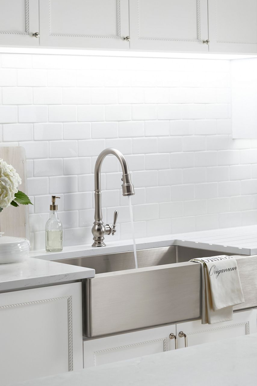 Angled view of a stainless steel farmhouse sink in a kitchen with white cabinets and a white tiled backsplash and a traditional stainless steel faucet. 