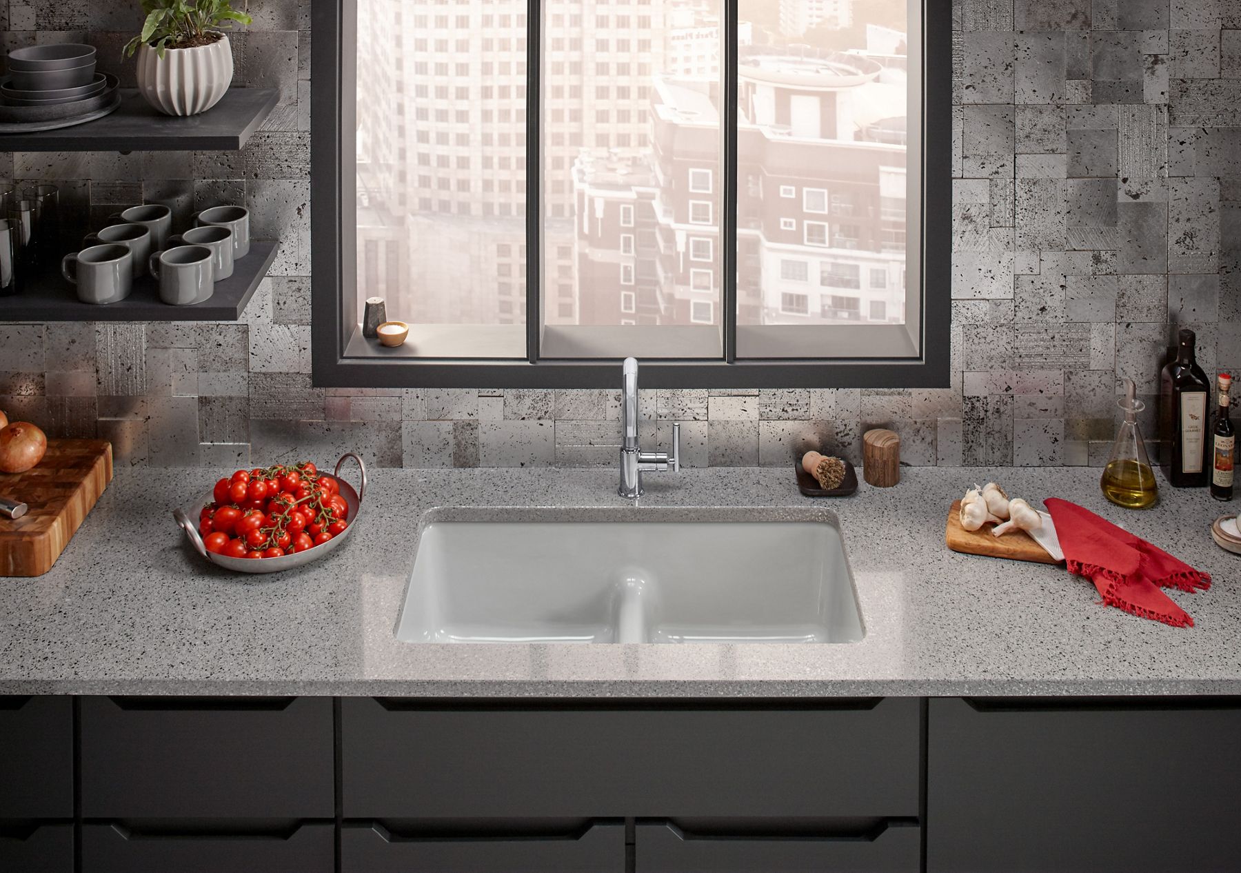 A kitchen counter with gray cabinets, a stone countertop, and smart divide sink, showing how the divider is lower than the top level of the sink, looking out from a high-rise window over city buildings below.