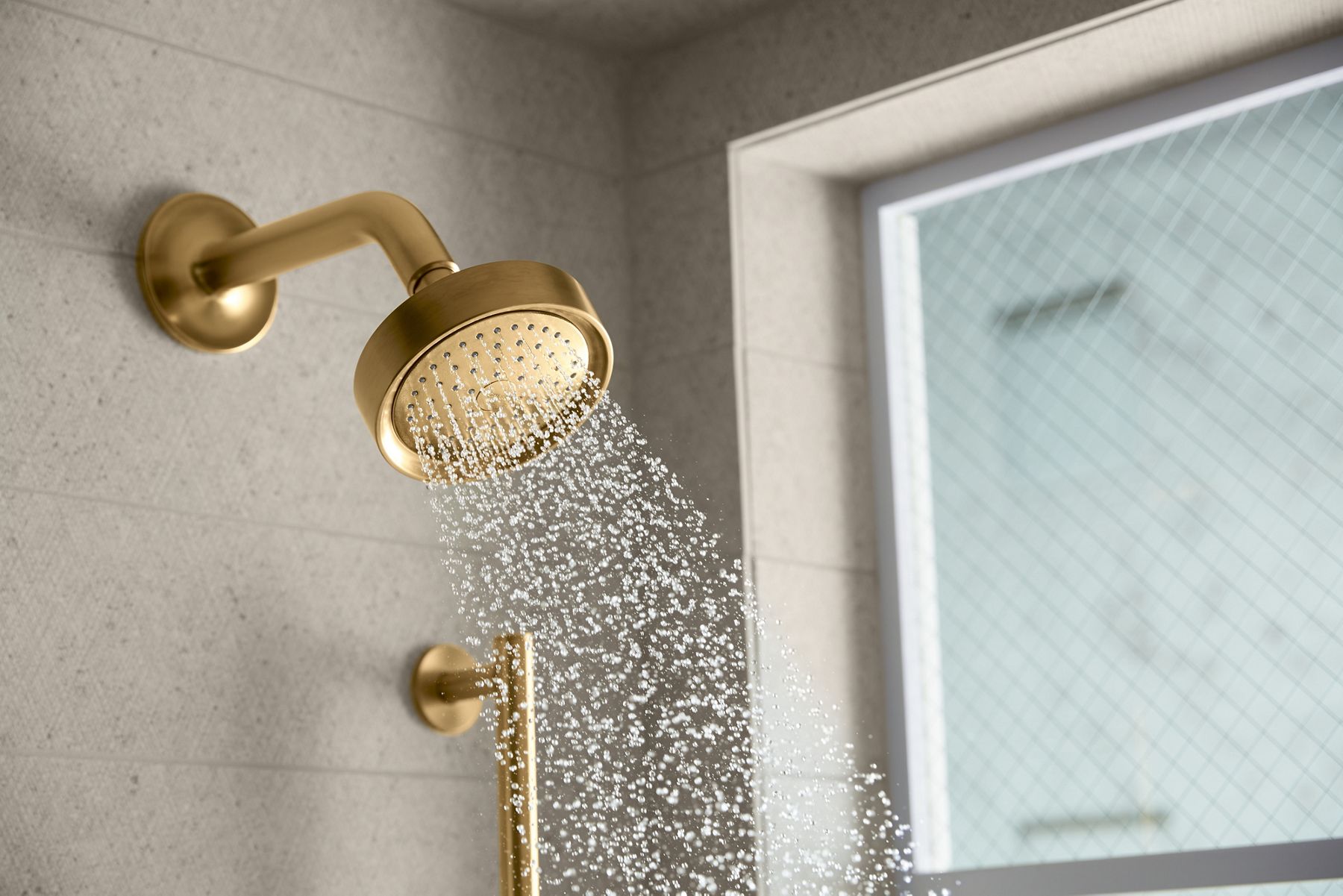 A brushed moderne brass shower head with water coming out of it and grab bar on a light gray tile wall.