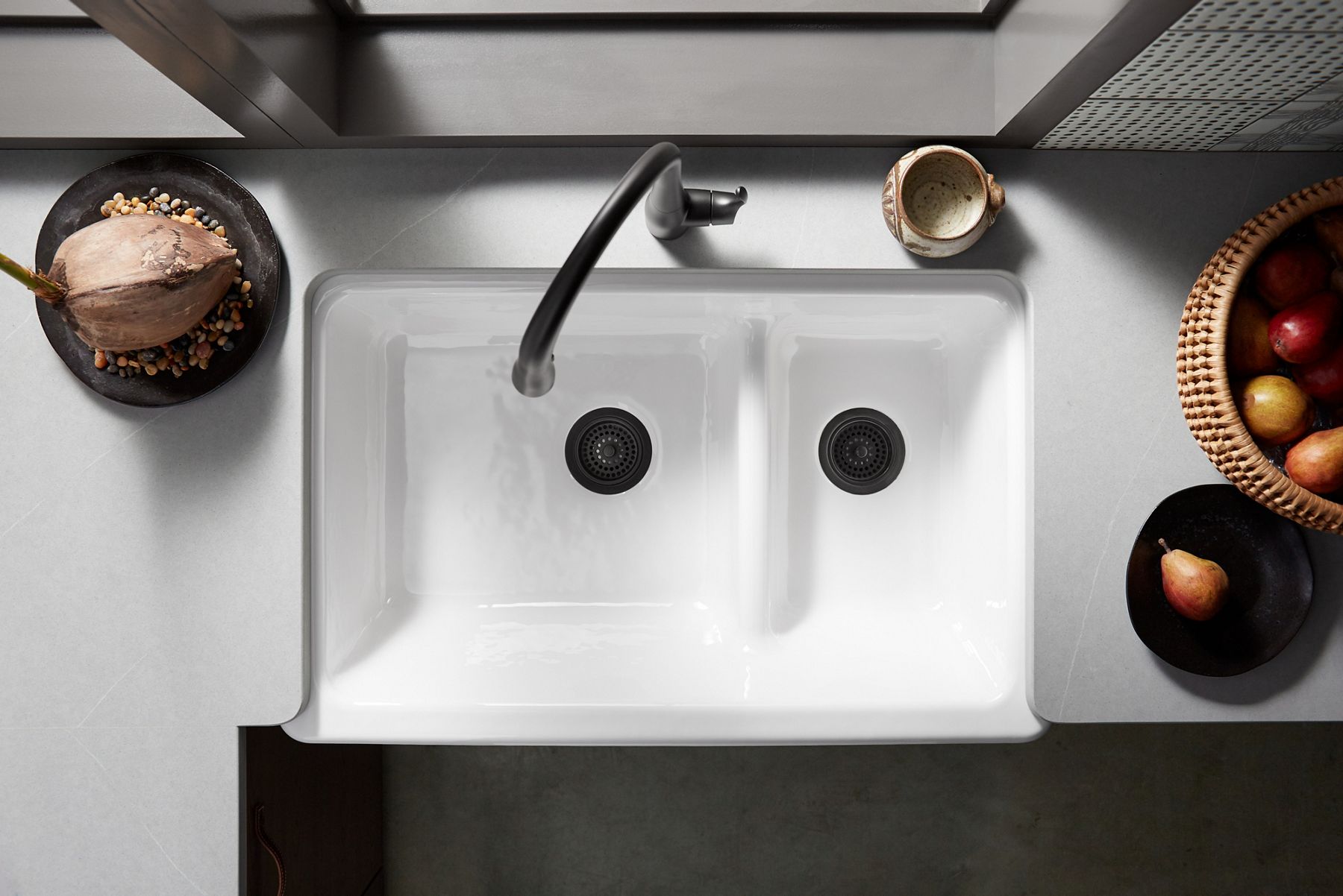 View from above of a wicker basket of apples beside a white KOHLER Whitehaven Smart Divide sink with a black Simplice single handle faucet.