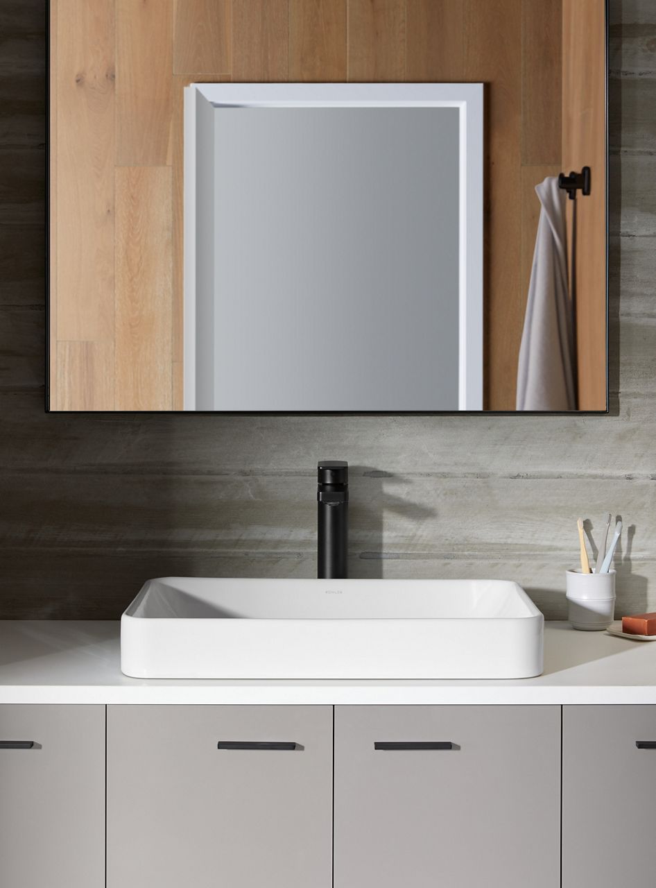 A contemporary bathroom with an oversized mirror, gray tile walls, white a Kohler Vox rectangle vessel sink, and Matte Black Parallel tall faucet.