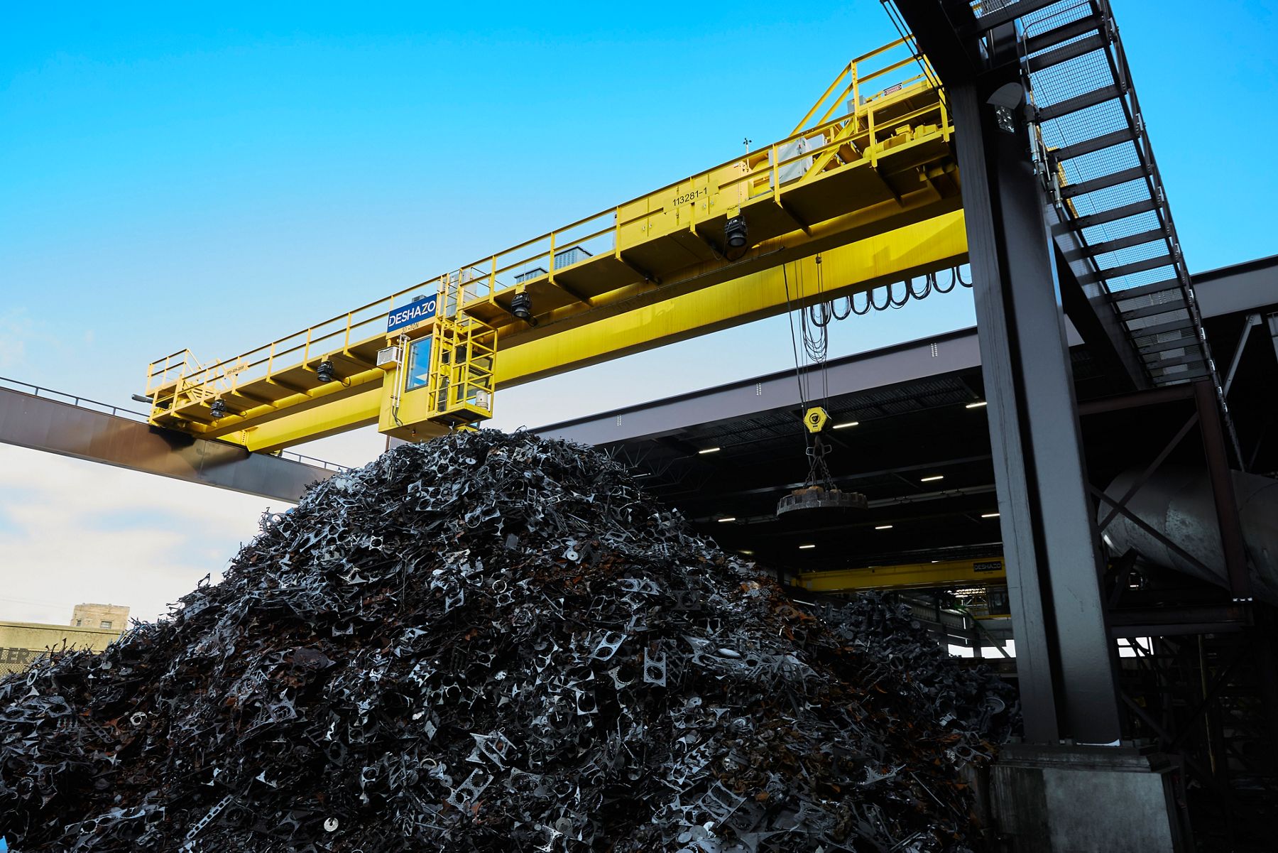 Large pile of recycled metal scrap beneath an overhead industrial crane inside a processing facility.