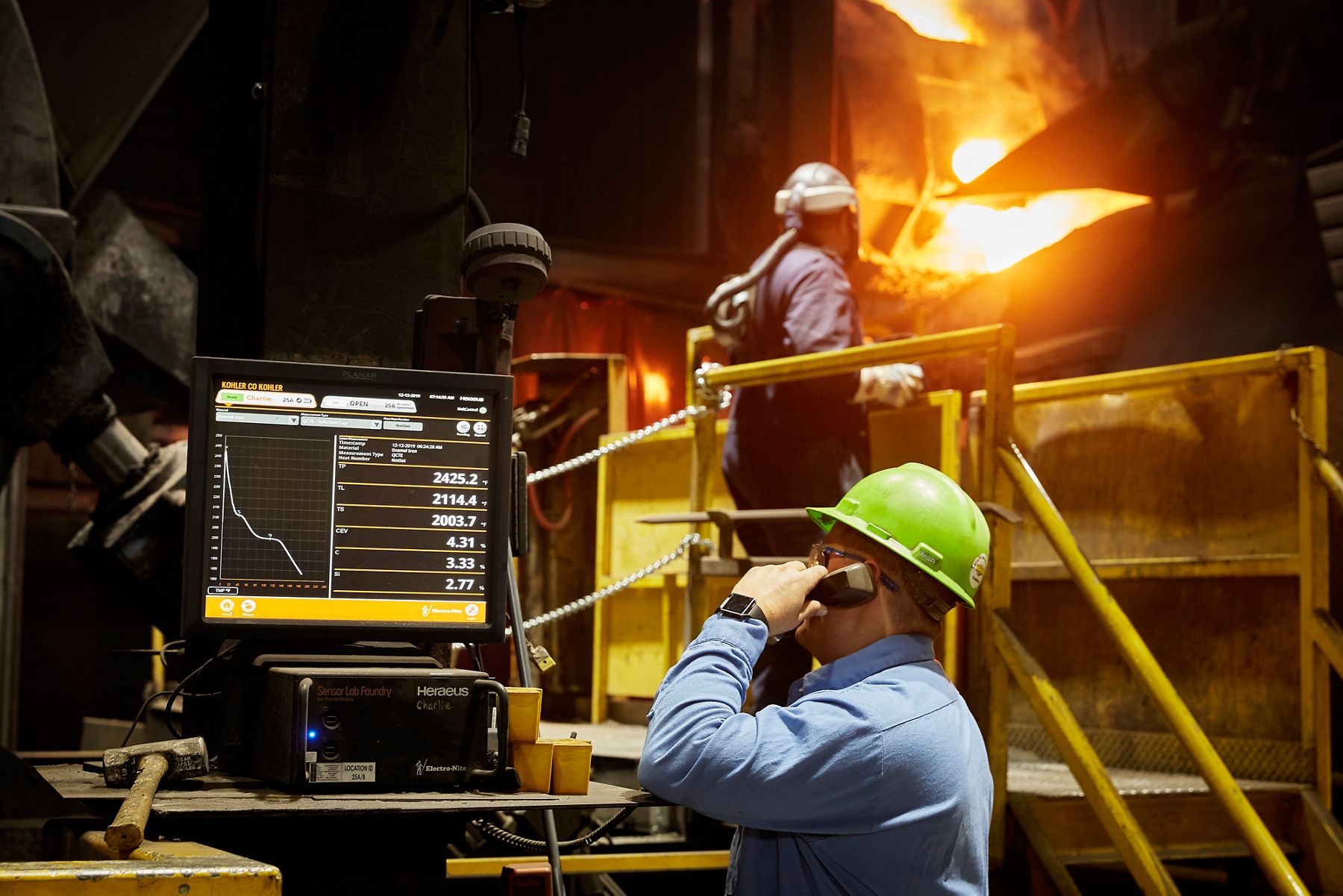 Industrial worker monitoring molten metal production with computer equipment inside a foundry.