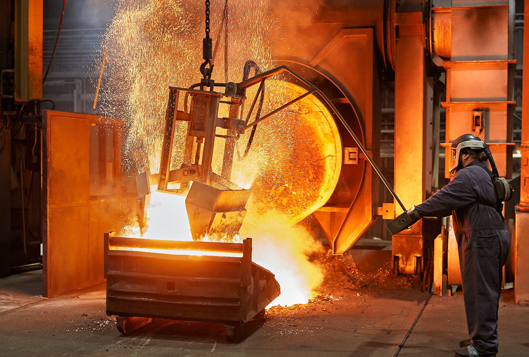 Foundry worker operating heavy equipment as molten metal pours and sparks fly inside an industrial facility.