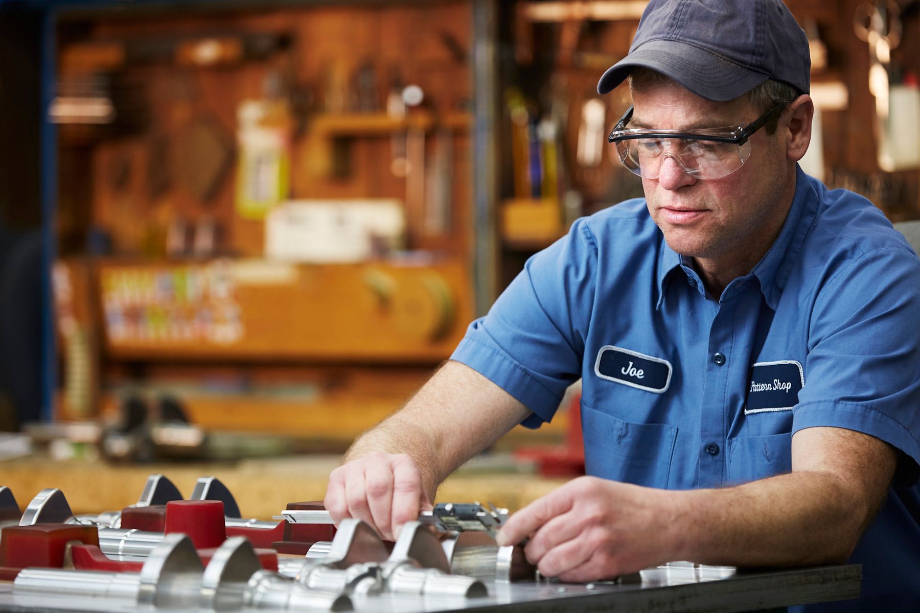 Factory worker in safety glasses inspecting machined metal components at a workbench.