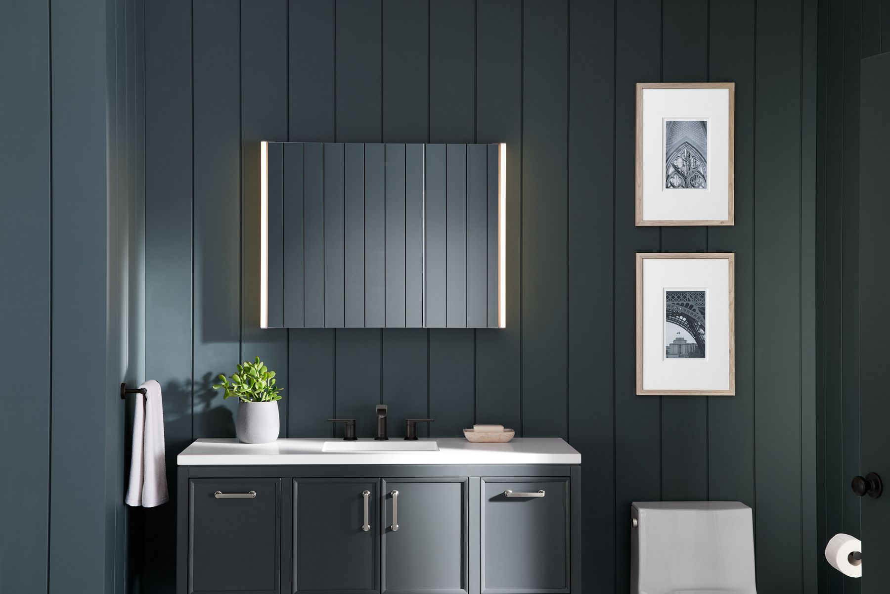 A KOHLER Verdera lighted medicine cabinet shines in a bathroom with dark gray walls, gray vanity, white vanity top, white toilet, and framed art with white mats.