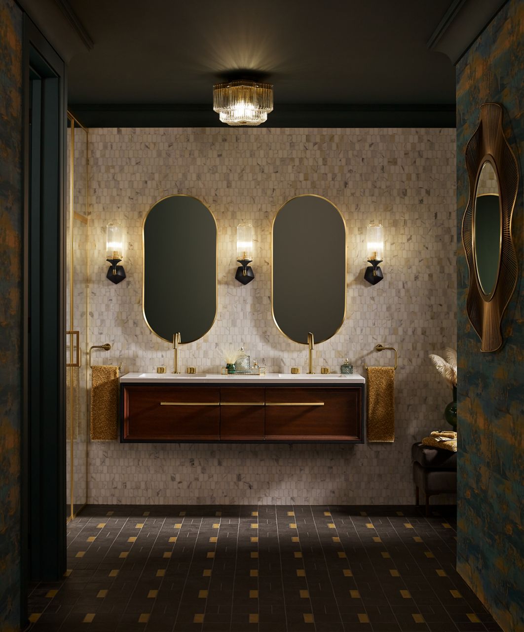 A bathroom styling space with a wall-mount double vanity in dark wood tones with brass accents and faucets, a multi-toned beige tile wall with brass-framed pill-shaped mirrors and two-toned sconce lights.