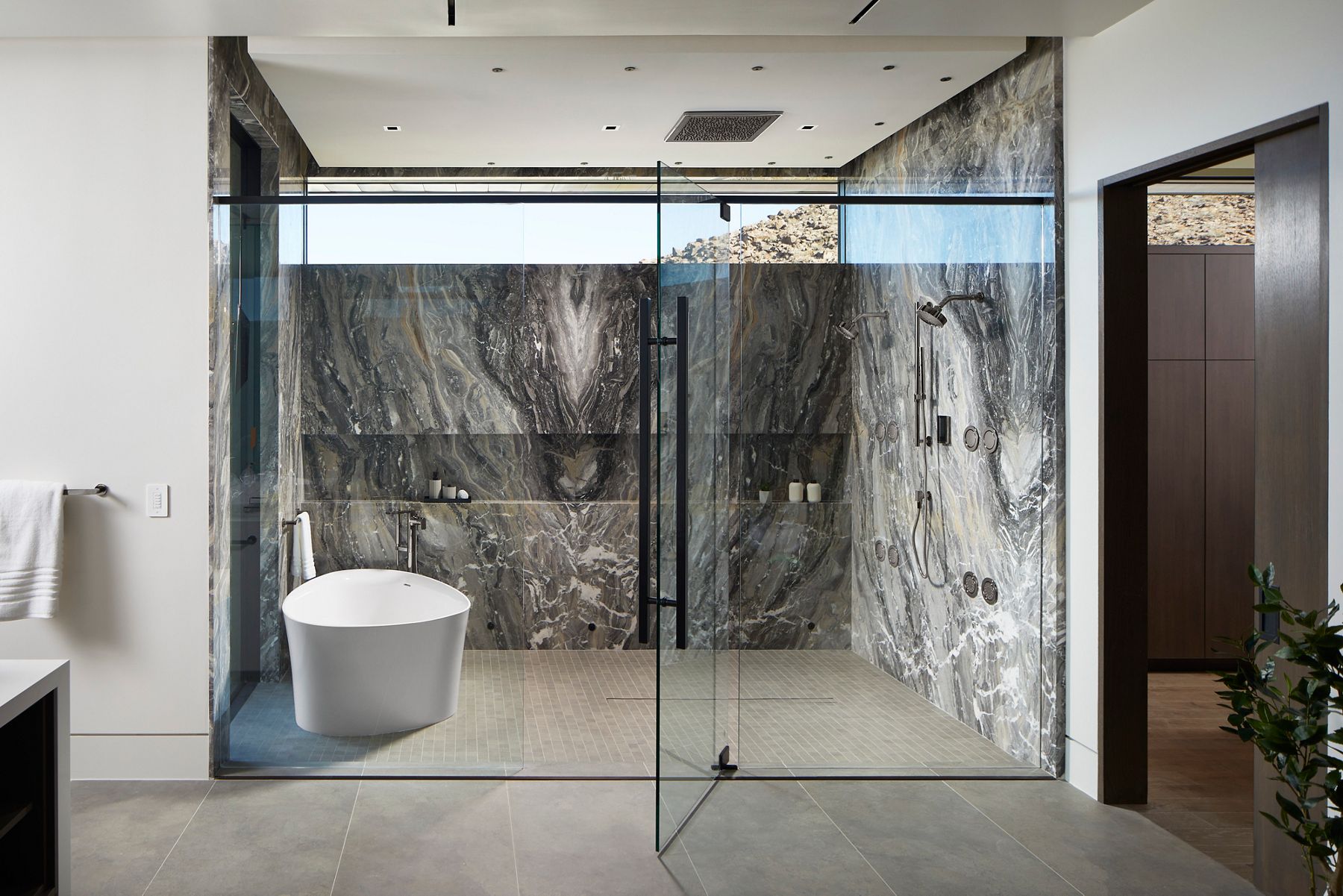 Modern and spacious shower with striking grey marble walls and floor. The glass enclosure showcases a minimalist design, with a ceiling-mounted square rain shower head and a sleek handheld shower wand. The space is well-lit, highlighting the marble's natural patterns.